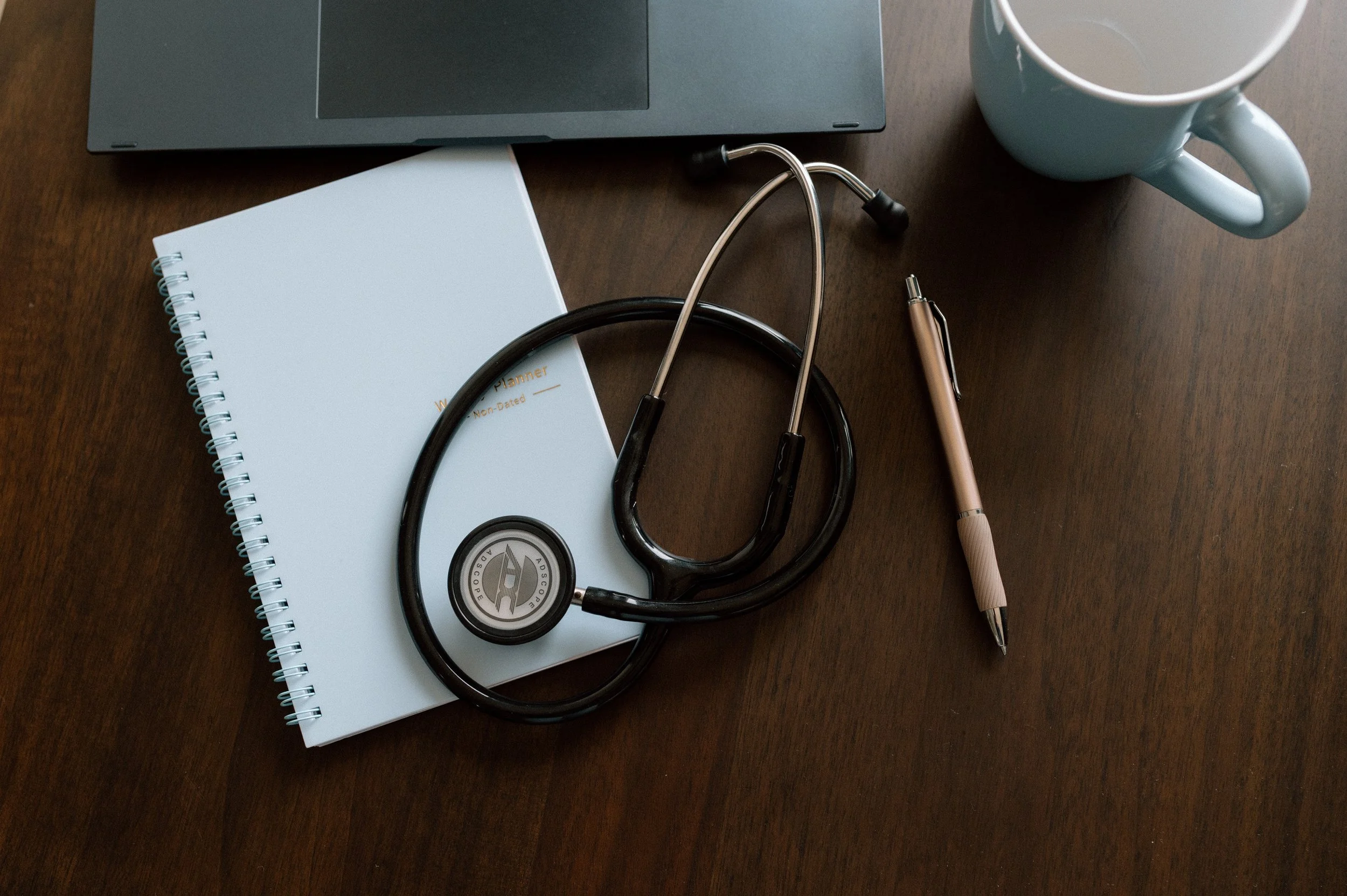 A stethoscope resting on a white notebook with a spiral binding, a silver pen, a laptop keyboard, a mug, and a pair of glasses on a wooden desk.