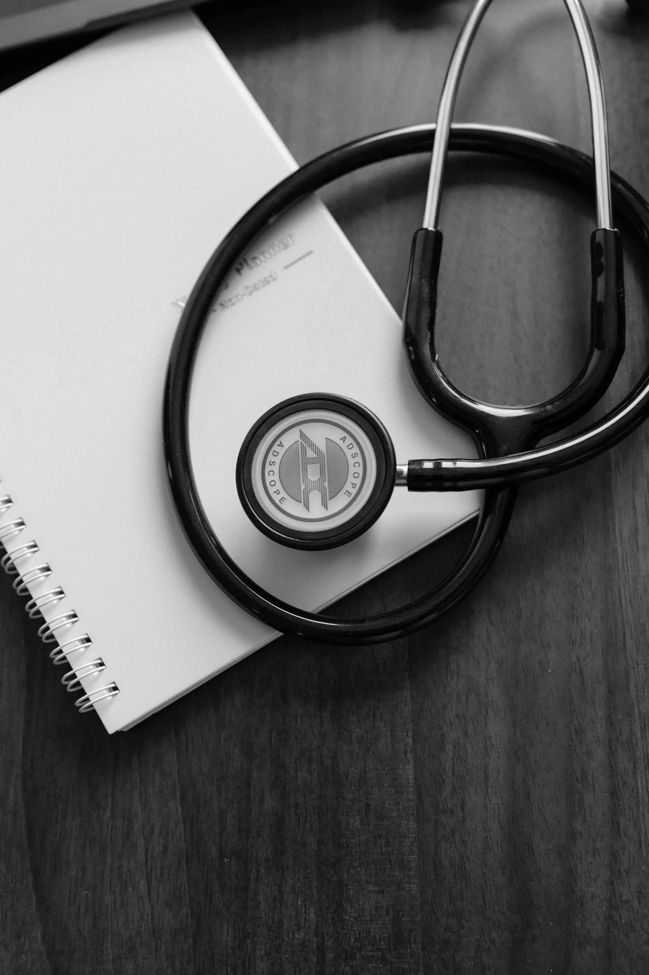 Black and white photo of a stethoscope resting on a closed notebook, placed on a wooden surface.