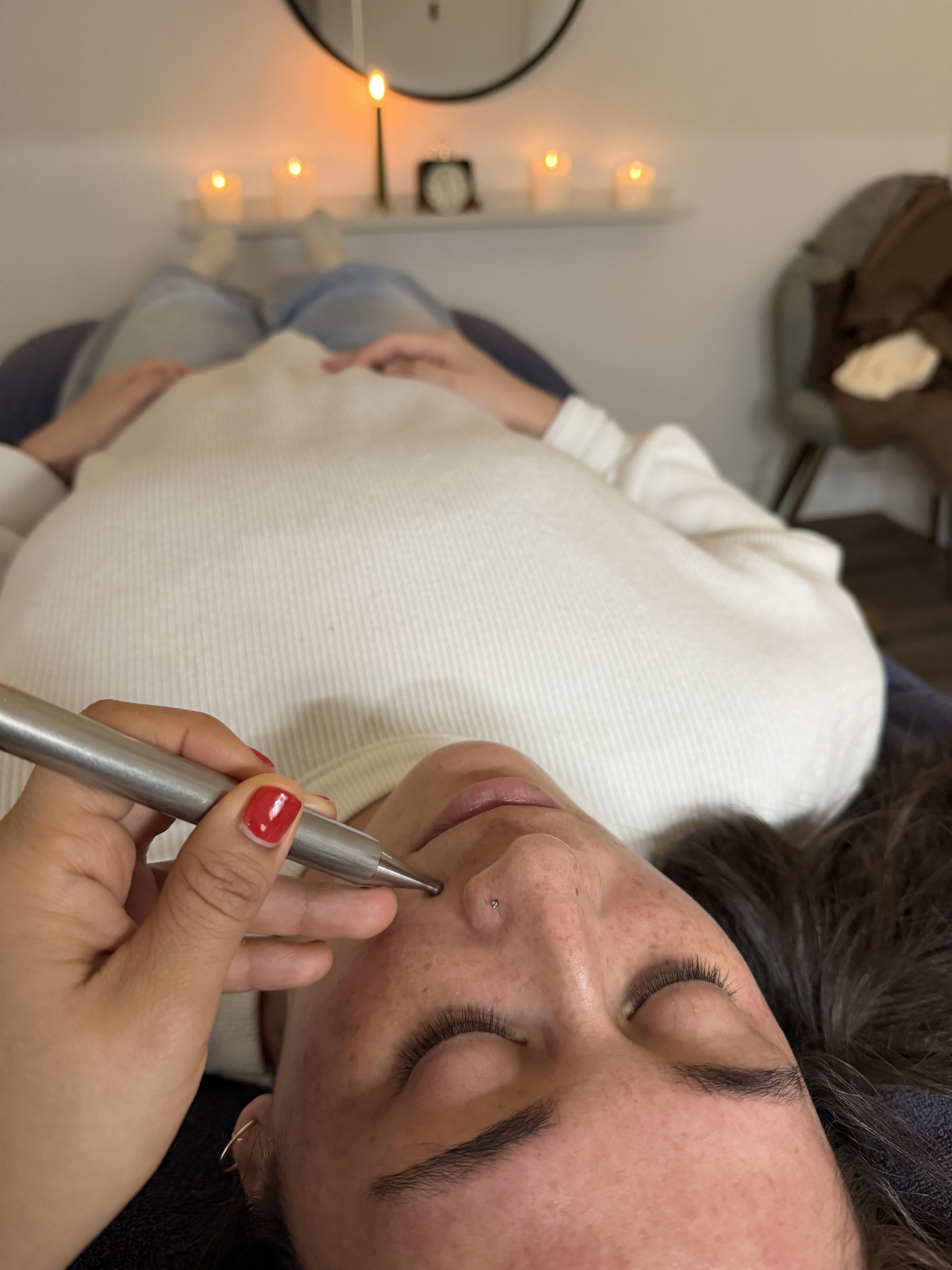 A woman with freckles and dark hair receiving a facial treatment using a pen-like device while lying down with eyes closed, on a bed with a person in the background, candles, and a mirror on the wall.