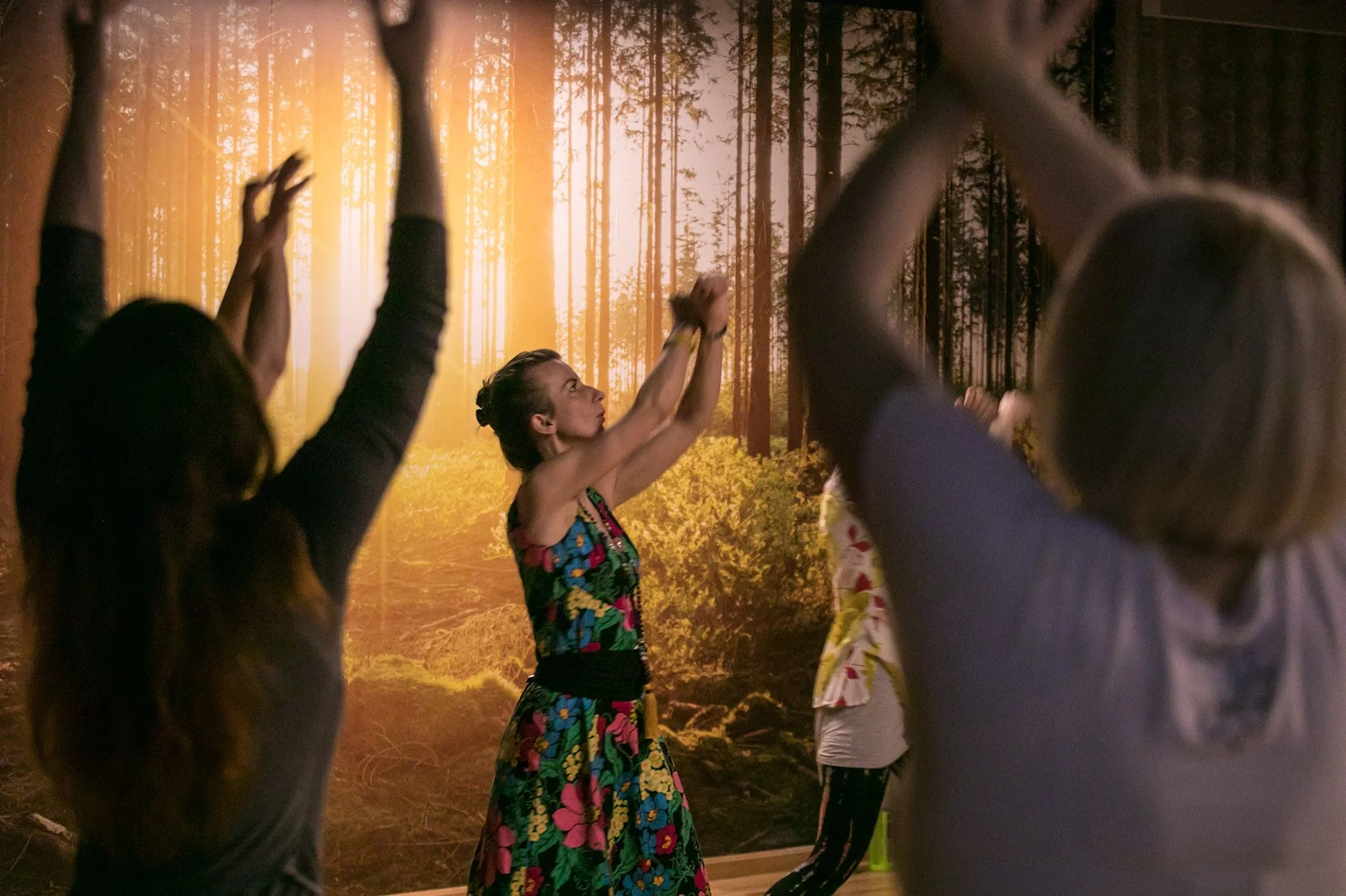 Inimesed Lomi-Lomi kursusel tegemas liikumisharjutusi, kõikide käed suunatud üles, ruumis, mille seinal on päikseline metsafoto.