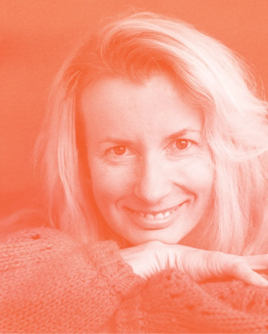 A woman with wavy blonde hair smiling, resting her chin on her hand, wearing an orange sweater, against an orange background.