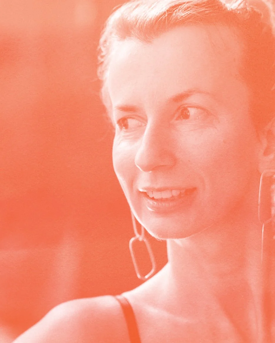 Close-up of a smiling woman with short hair, wearing earrings and a tank top, against a blurred background with a reddish filter.
