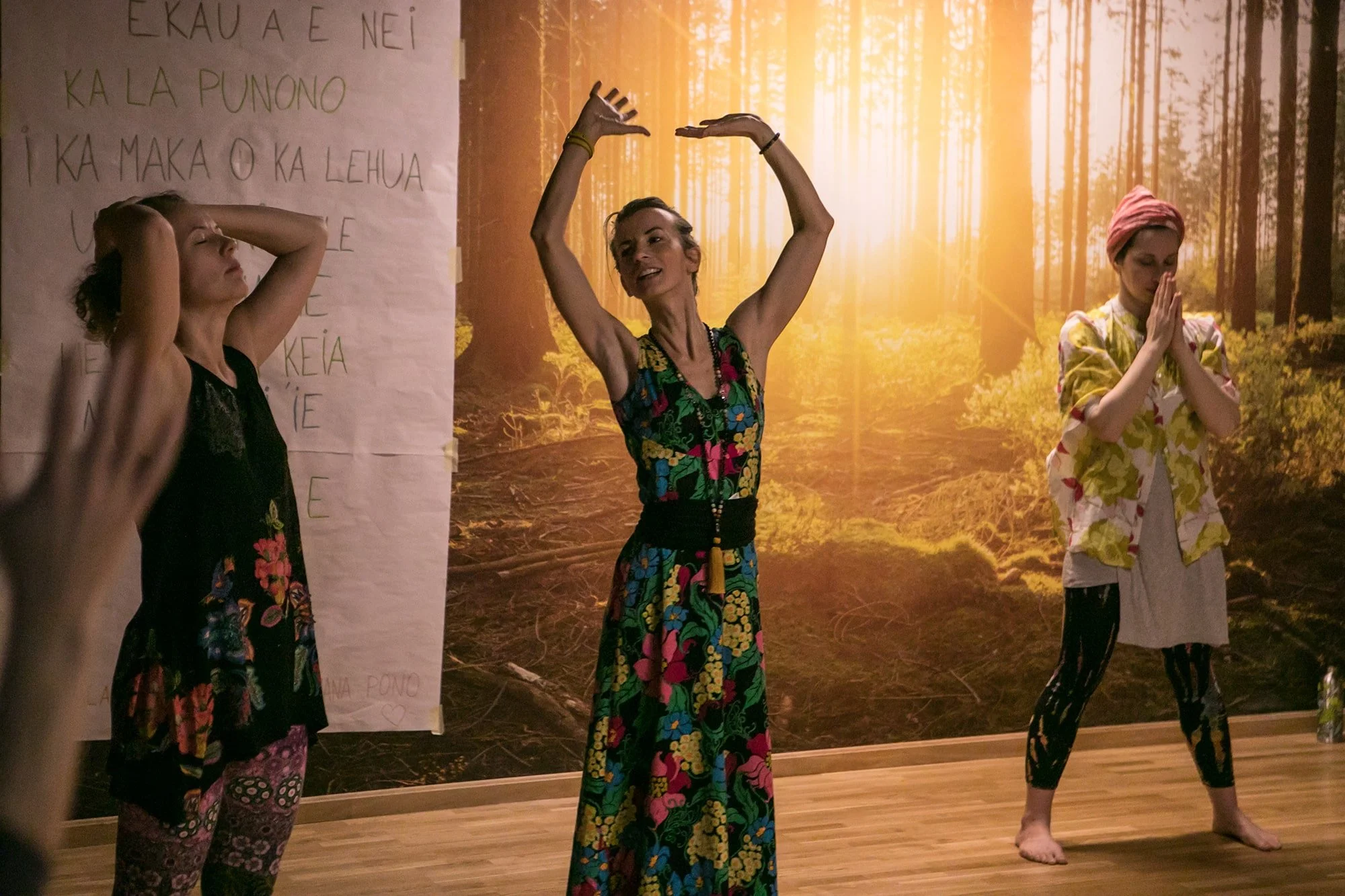 Riita juhendamas Lomi-Lomi koolitust, käed üleval, tegemas erinevaid liikumisharjutusi, taustaseinal on päikseline metsafoto.