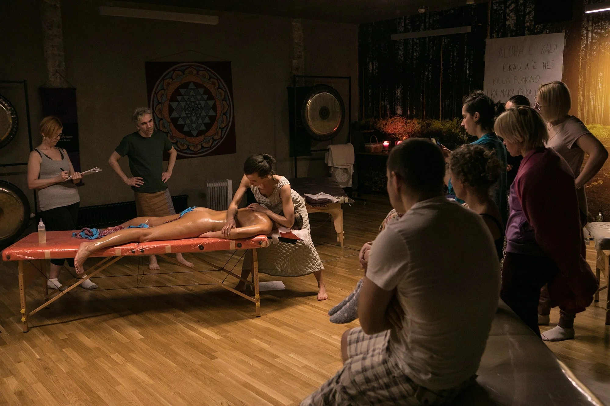 A group of people observing a massage training session on a massage table in a room with wooden floors, surrounded by wall decor and musical instruments.