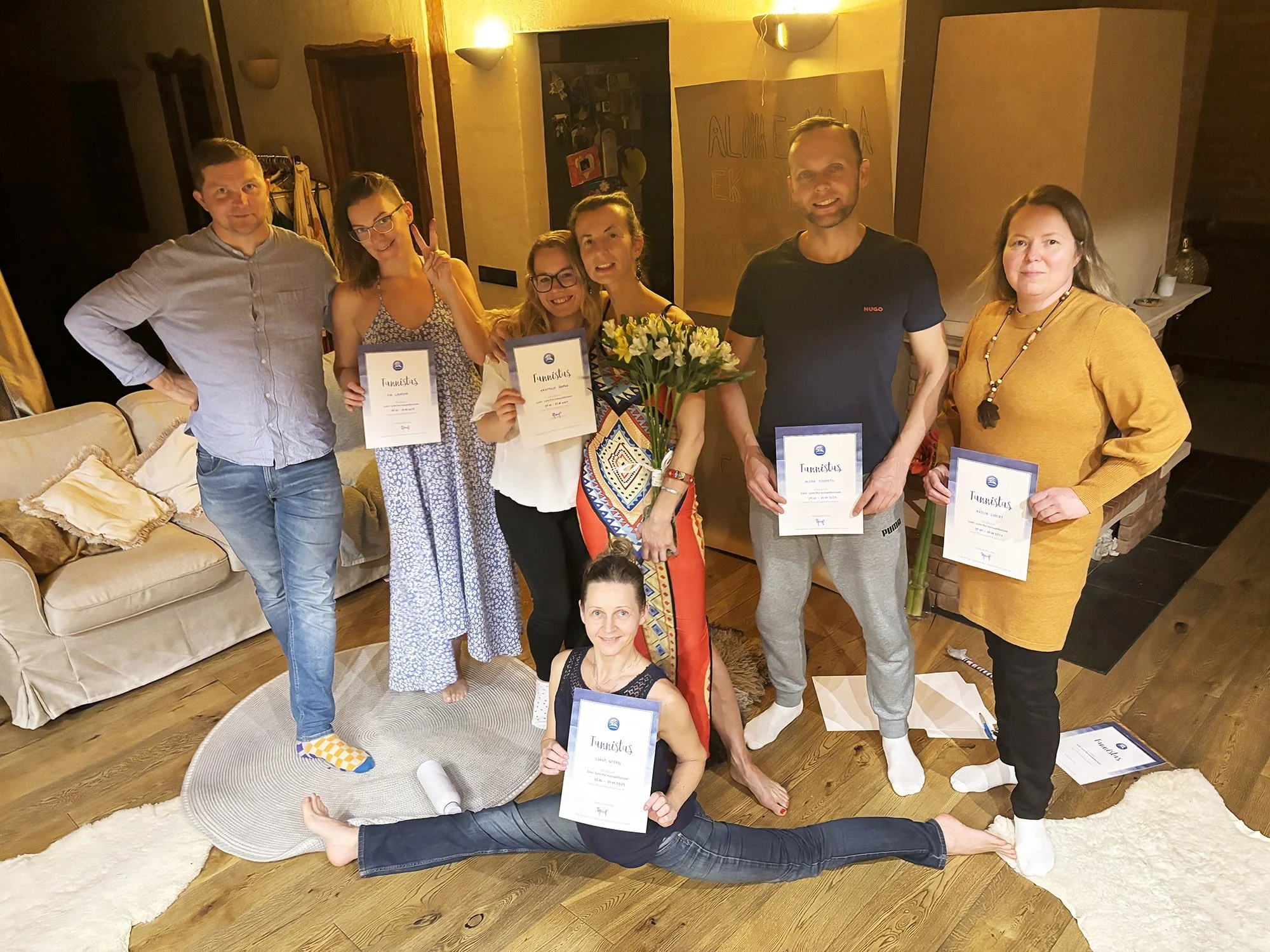 Seitse inimese rühm hubases ruumis hoiab käes tunnistusi. Üks inimene istub põrandal, tehes spagaati ja hoides tunnistust, keskel on Lomi-Lomi koolitaja Riita, kes hoiab käes lillekimpu. Nad naeratavad ja poseerivad foto jaoks.