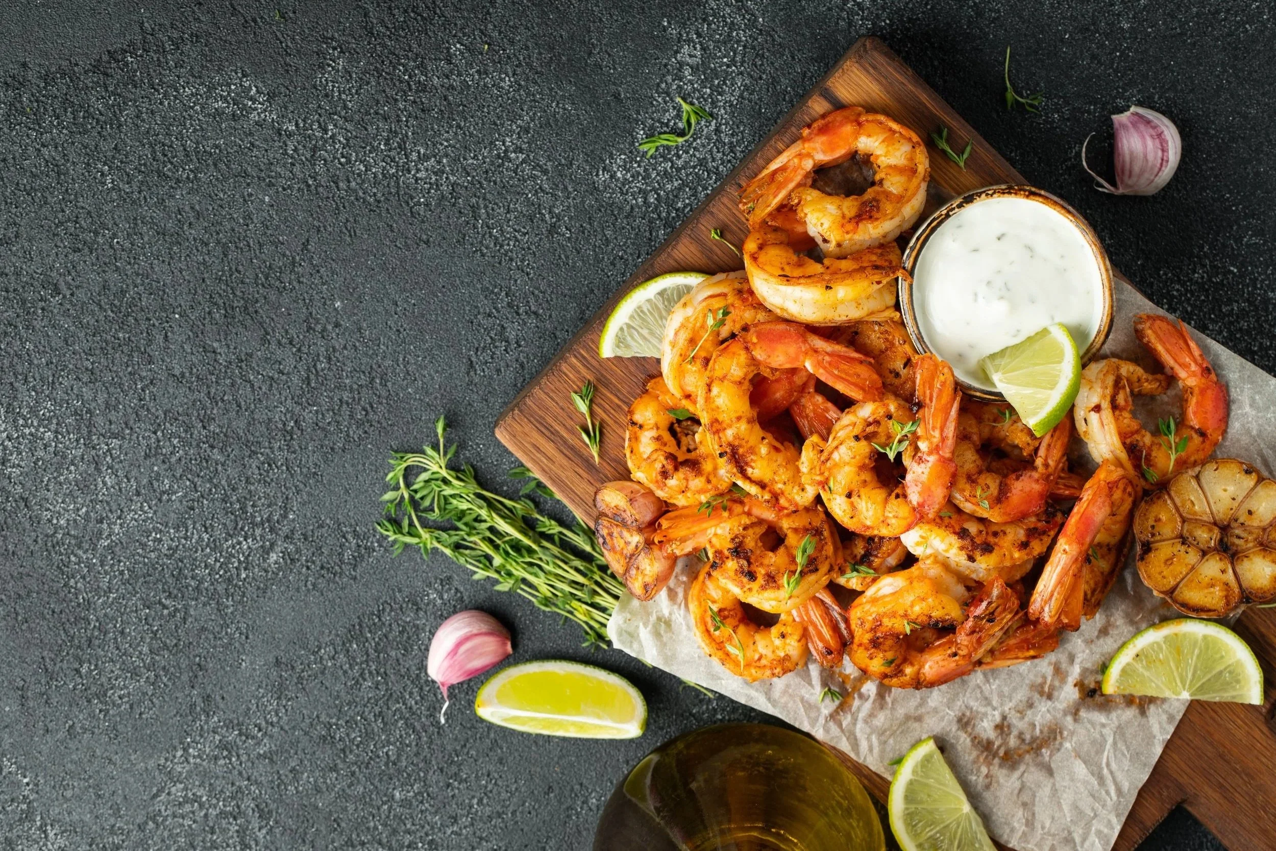 Shrimp on a wooden platter with lemon wedges, garlic, herbs, and a bowl of dipping sauce, on a dark textured surface.