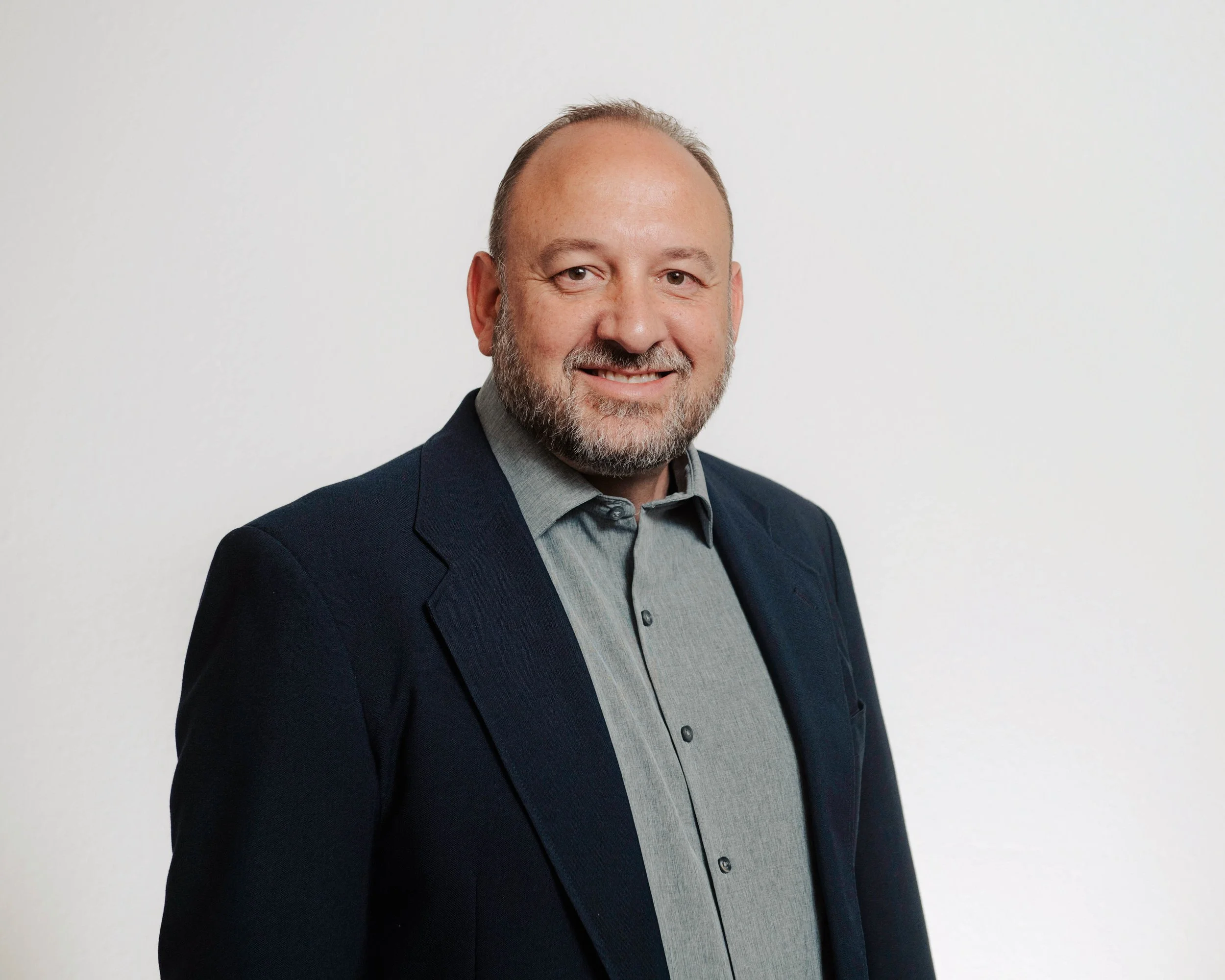 Headshot of a smiling middle-aged man with a beard, wearing a gray shirt and navy blazer, standing against a plain white background.
