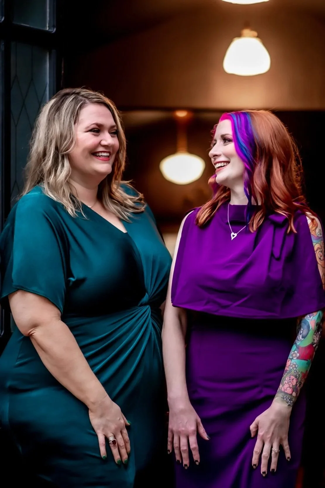 Shylah Ridgway and Dana Kausek of Ala Therapy Collective, two women smiling at each other indoors. One with blonde hair in a teal dress, the other with red and purple hair in a purple dress with arm tattoos. Affirming online therapy in Oklahoma.
