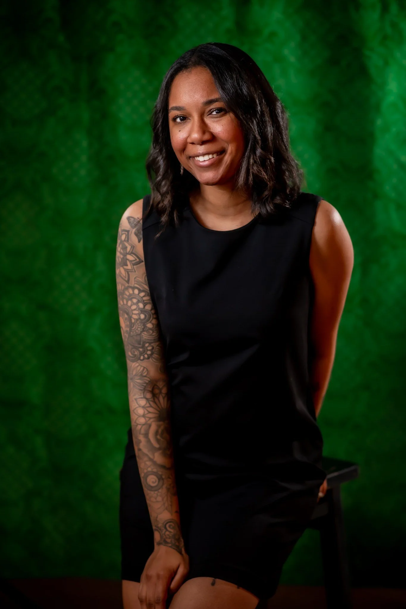A smiling woman with shoulder-length dark hair wearing a black sleeveless dress and showing a tattooed arm standing in front of a green textured background.