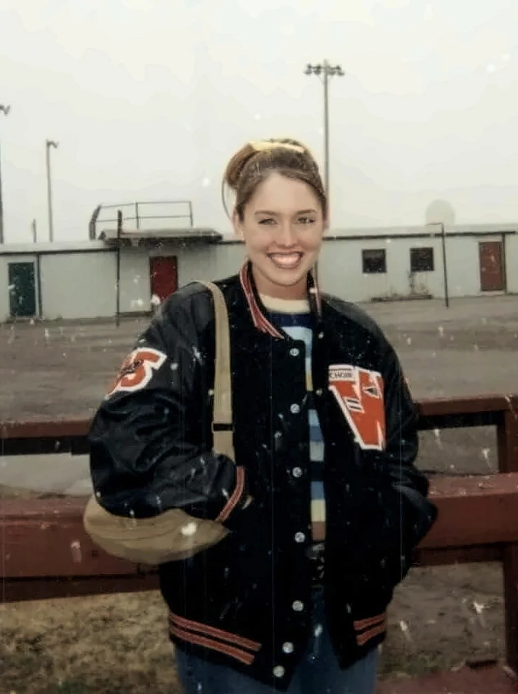 Teenage girl in a black and orange letterman jacket smiling outdoors in falling snow, standing by a school athletic field