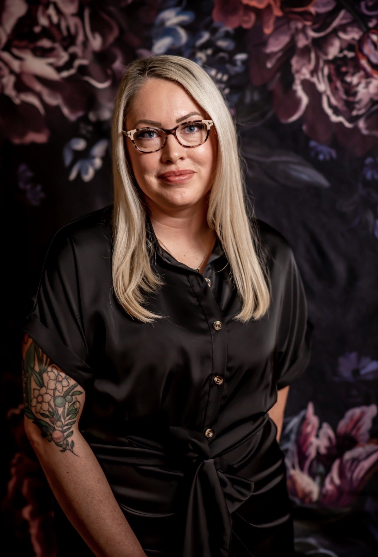 Taylor Hendricks, LMSW, ADHD and autism affirming therapist at Ala Therapy Collective, standing confidently in a black button up against a dark floral background with a floral tattoo on her left arm.