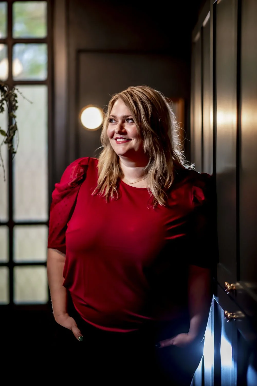 A smiling woman with shoulder-length blonde hair wearing a red top, standing indoors near a dark wall and window, with soft lighting in the background.