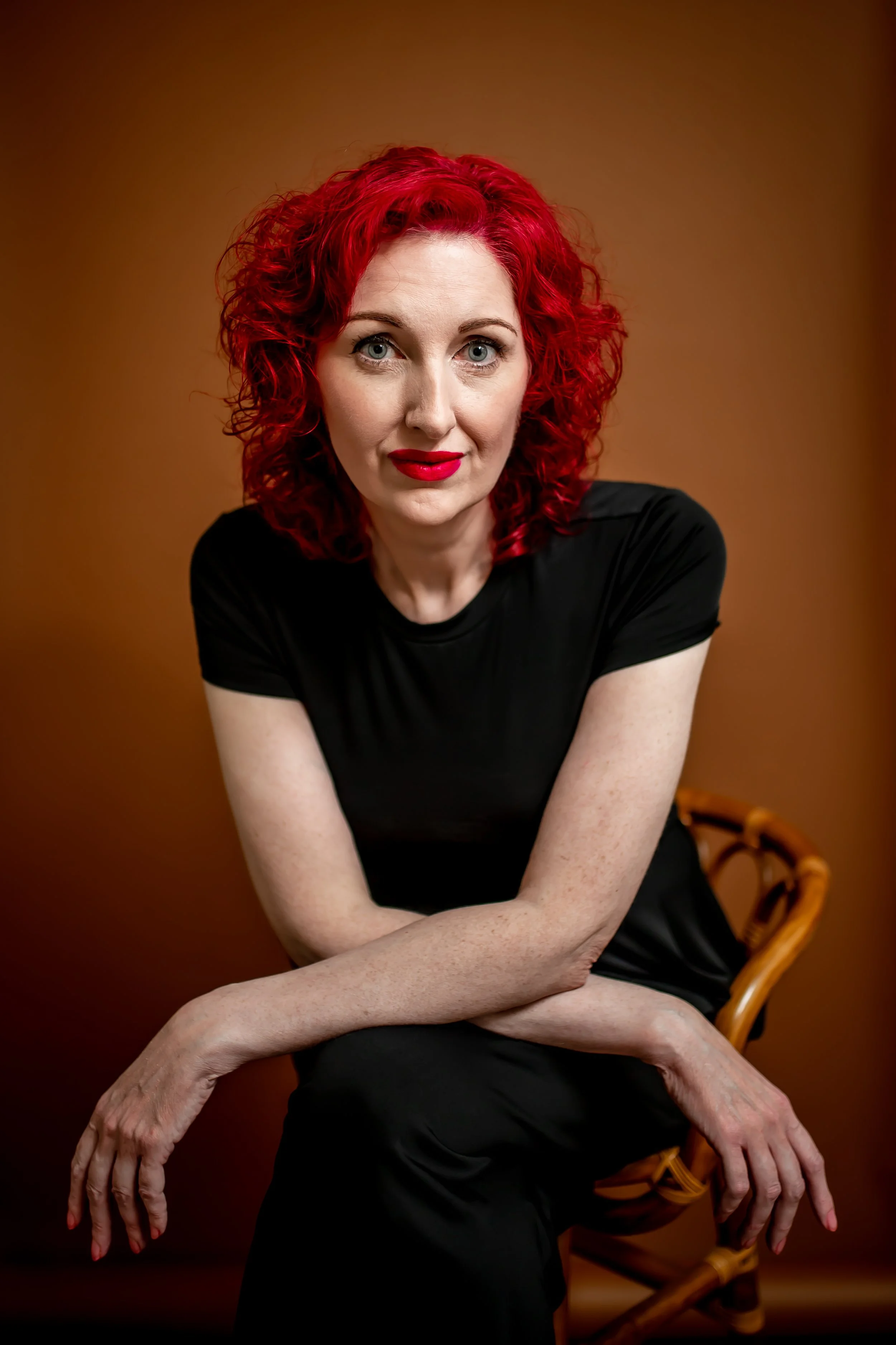 Michi Medley, LMSW, A-CAS of Ala Therapy Collective; a woman with vibrant red curly hair in a black t-shirt, sitting on a wicker chair. Specialist in neurodiversity, ADHD, and autism-affirming therapy in Oklahoma.