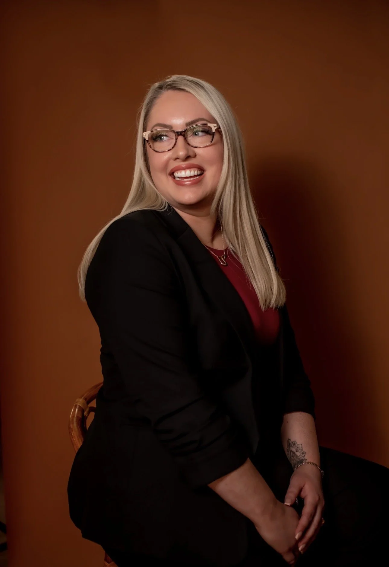 Taylor Hendricks, LMSW, online therapist for women and LGBTQIA+ clients in Oklahoma, laughing warmly in a black blazer and red top, standing by a wooden chair against a brown background.