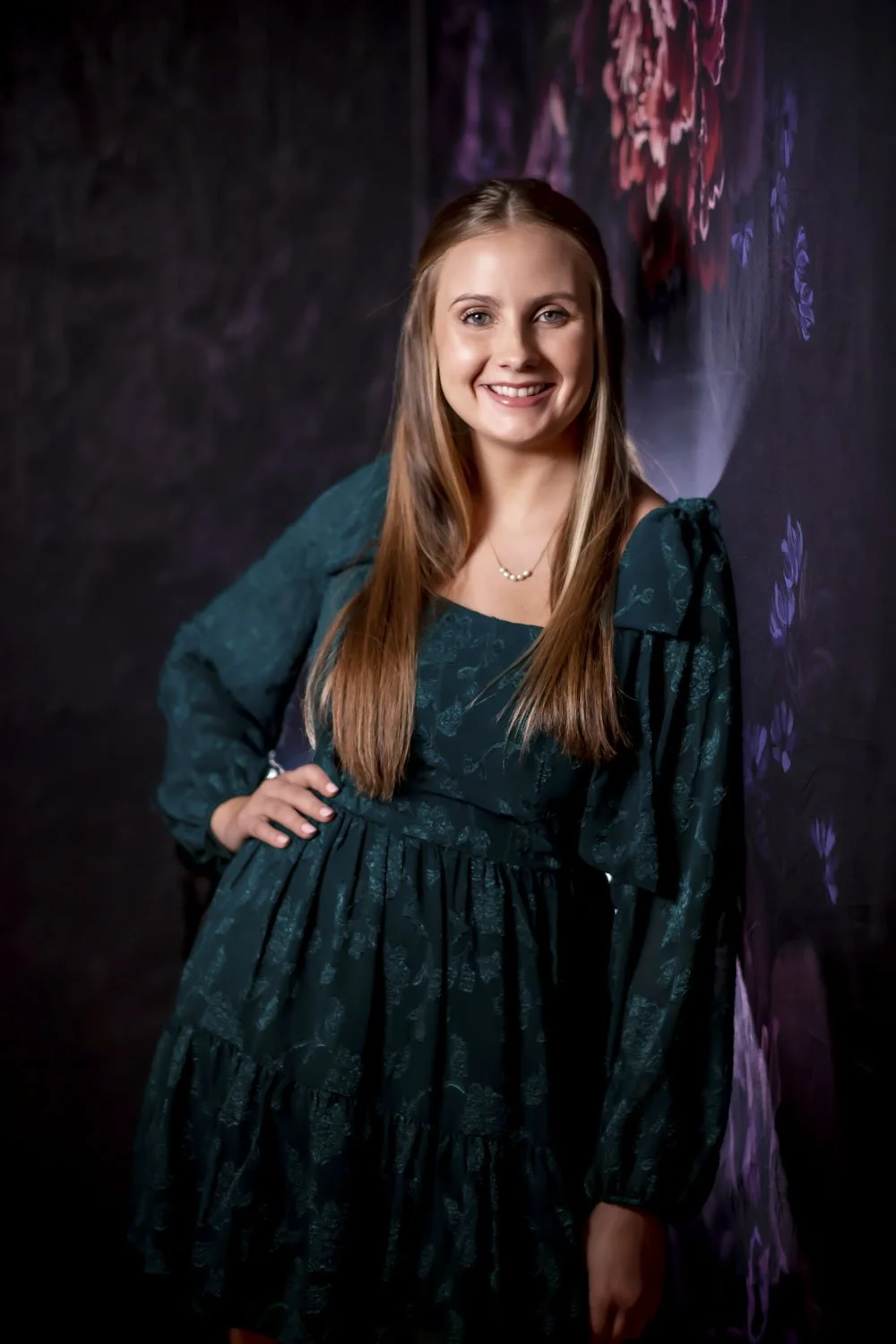 A young woman with long, light brown hair standing against a dark, floral or painted backdrop. She is smiling, wearing a dark green dress with long sleeves and a small pearl necklace.