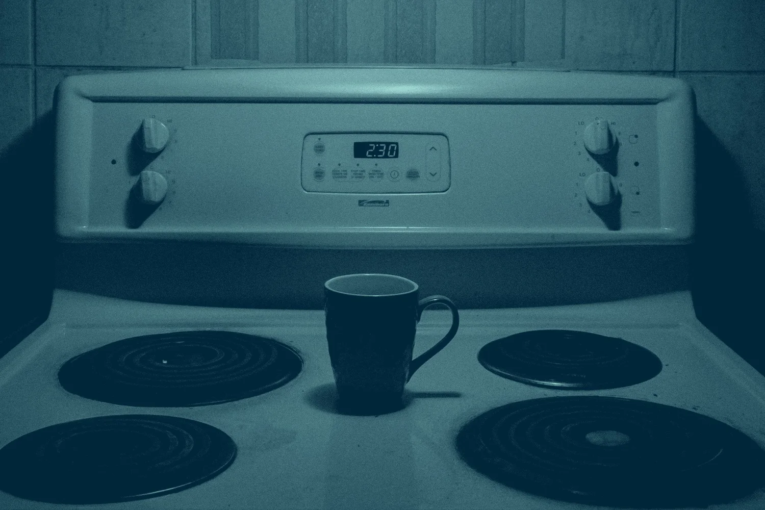 A kitchen stove clock reading 2:30 in the morning beside a single mug on a dark cooktop
