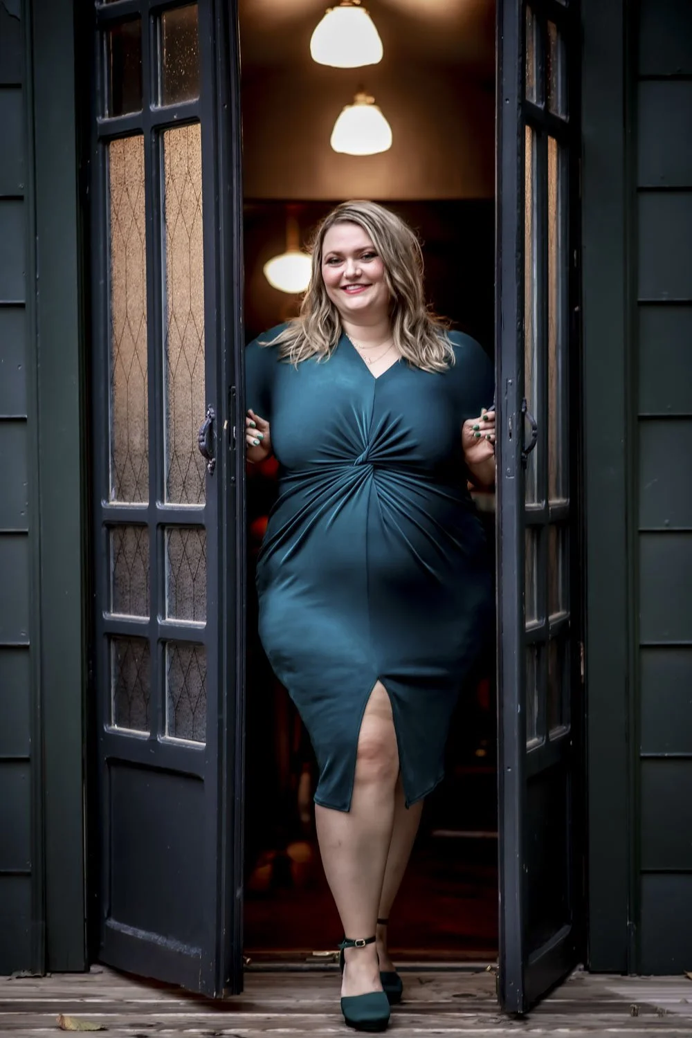 A smiling woman in a teal dress with a slit, standing at a doorway with open glass-paneled doors, inside a warmly lit room.