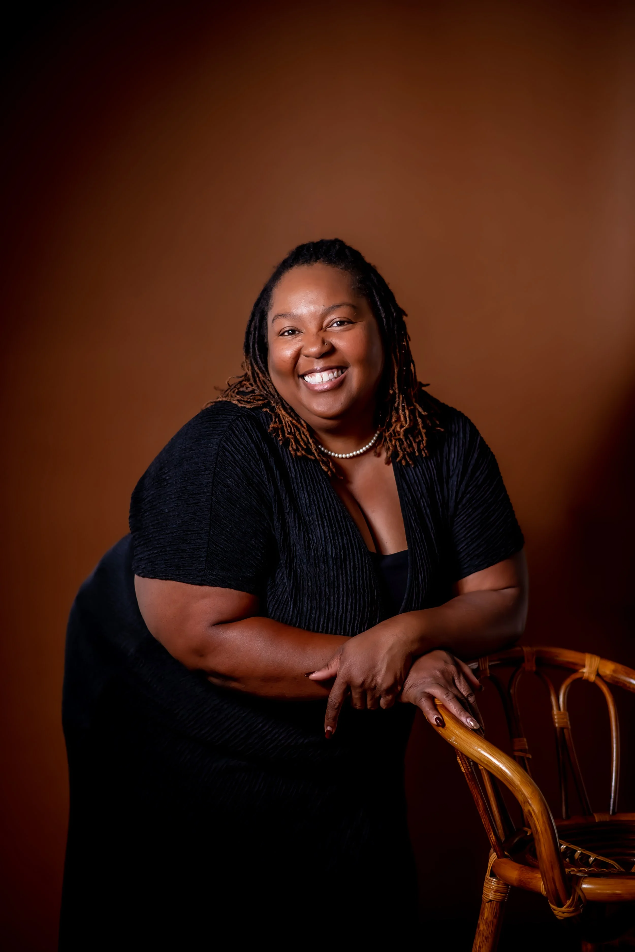 Demetria Bonds, LMSW of Ala Therapy Collective; a smiling woman with dreadlocks in a black dress and pearl necklace, leaning on a wooden chair. Providing inclusive online therapy for marginalized and rural communities in Oklahoma.