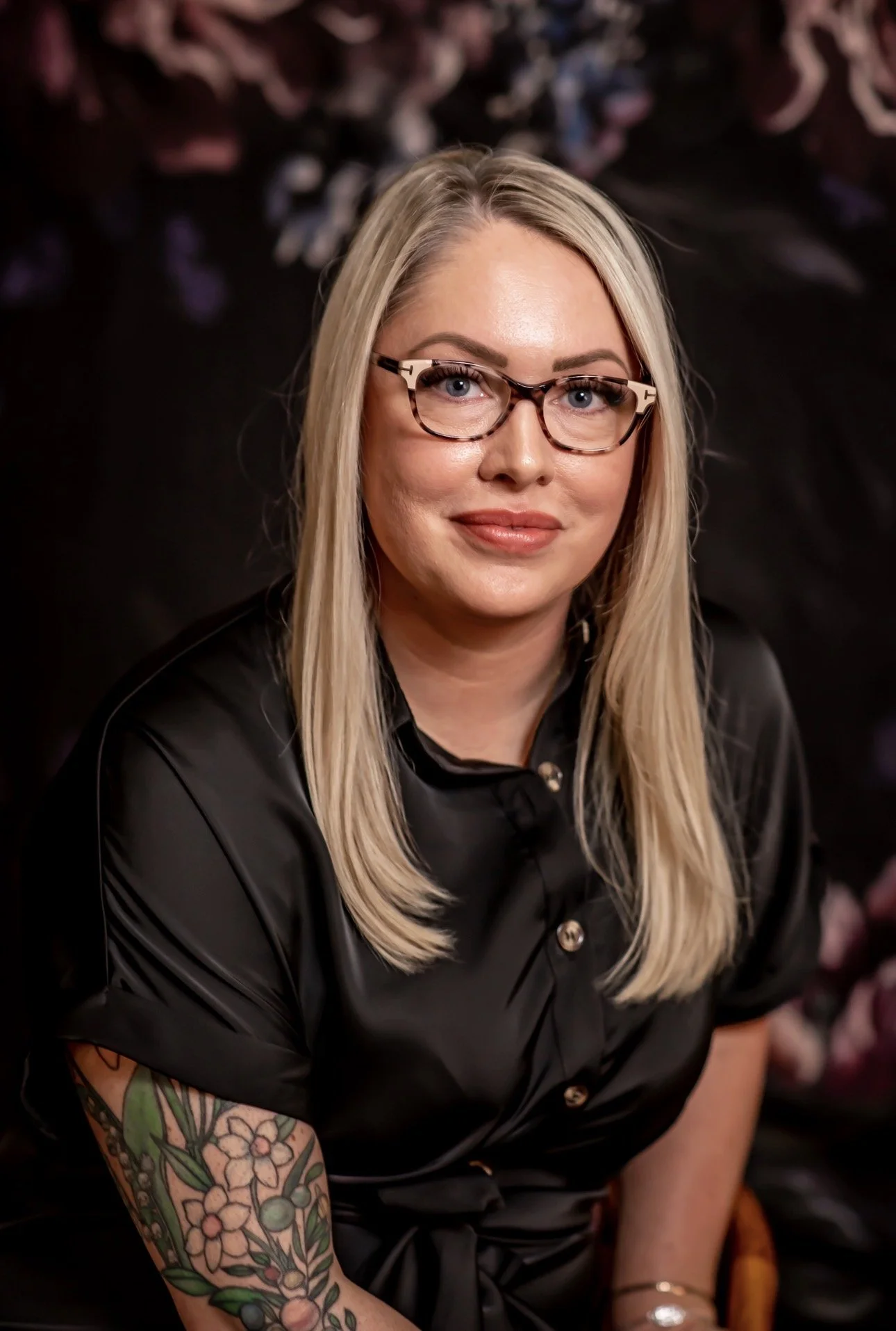 Taylor Hendricks, LMSW, neurodivergent affirming therapist in Oklahoma, smiling softly in a black silk shirt against a moody floral backdrop and showcasing a colorful floral tattoo on her left arm.