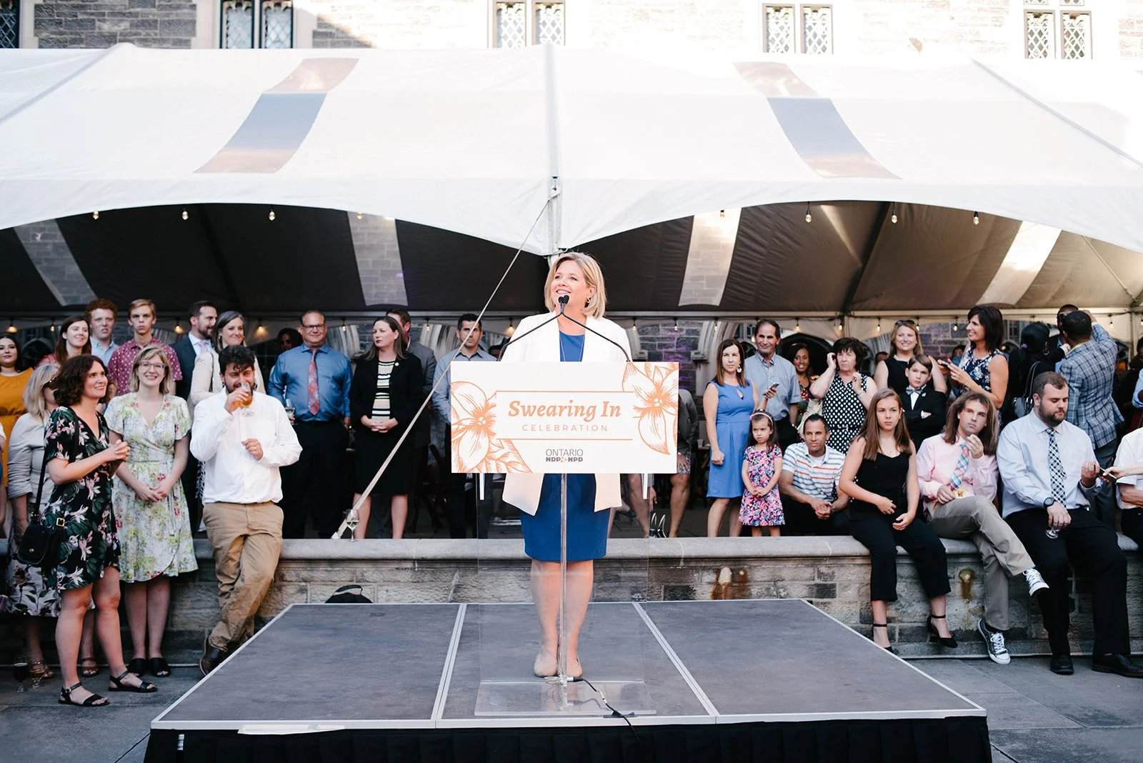Andrea Horvath in a blue dress and white blazer speaking at a podium with a sign that reads 'Swearing-In Celebration' in front of a large crowd of people under a white tent, outside a brick building.