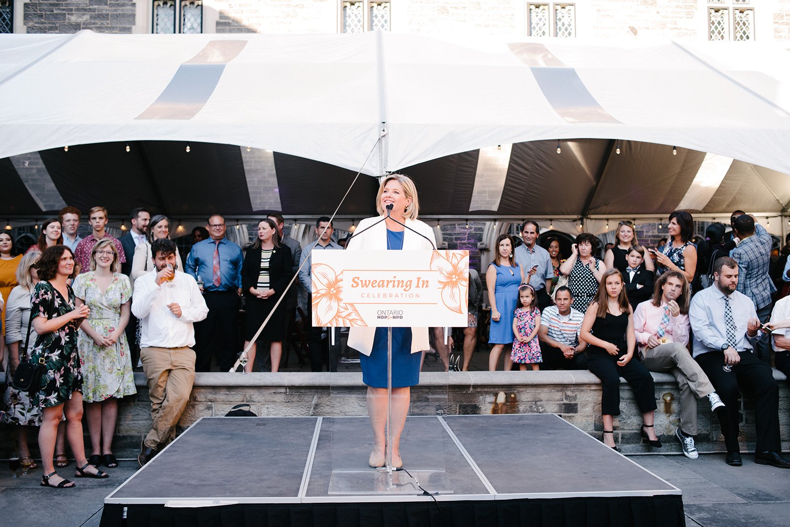 Andrea Horvath giving a speech at a podium at Hart House during an NDP swearing-in celebration under a large white tent, with a diverse group of people standing behind her.