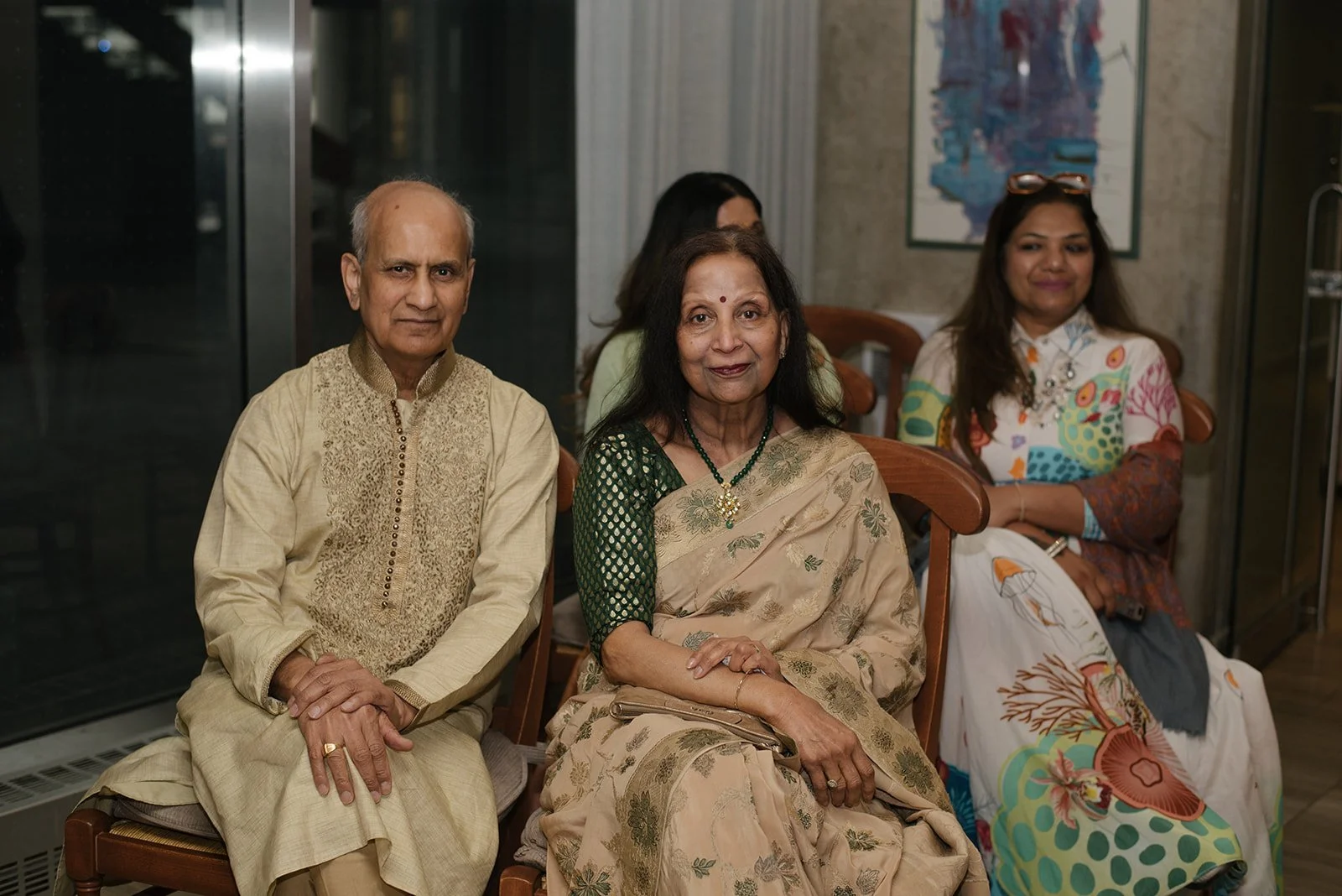 Four people sitting on chairs in a room with a glass door and artwork on the wall, two women and two men, dressed in traditional Indian attire.