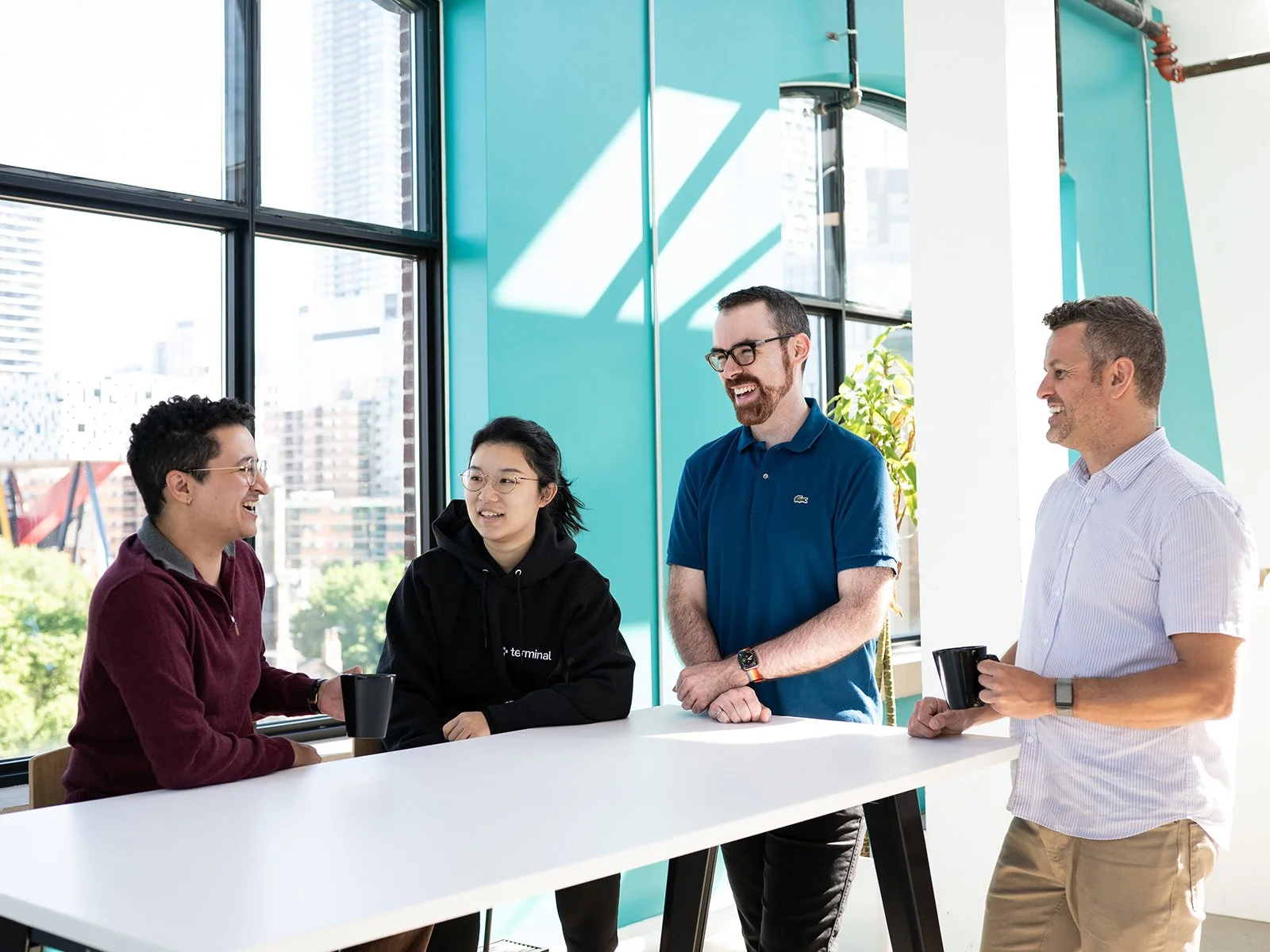 Four employees having a conversation in a bright modern office with large windows, sunlight, and cityscape view.