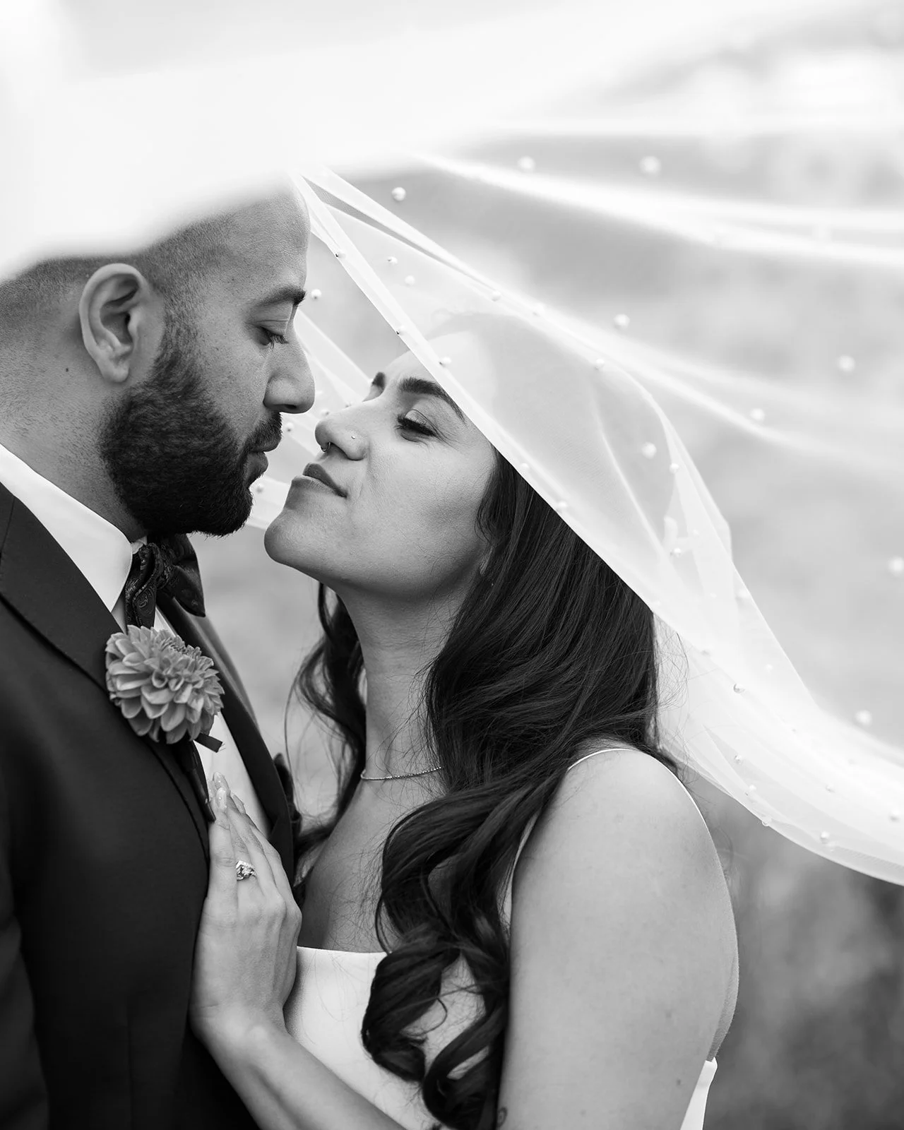 Black and white photo of a couple on their wedding day, standing closely with faces almost touching, under a sheer veil with pearl embellishments. The man has a beard and is wearing a suit with a bow tie, and the woman has long wavy hair, a ring visible on her finger, and is wearing a sleeveless dress.