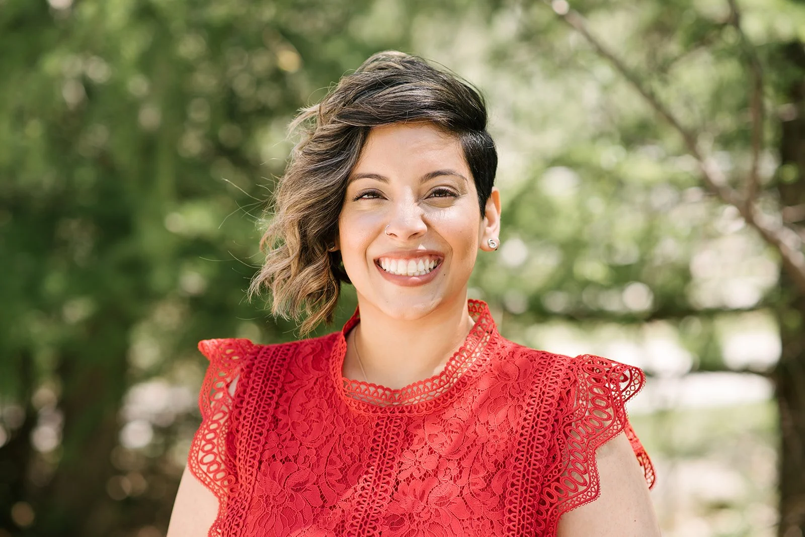 A headshot of a smiling woman with short dark hair wearing a red lace dress outdoors with green trees in the background.