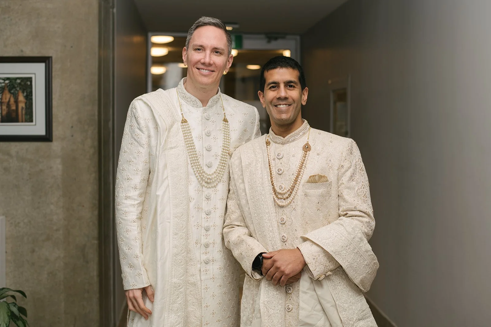 Two grooms in traditional Indian wedding attire standing indoors, smiling at the camera outside of Toronto City Hall.