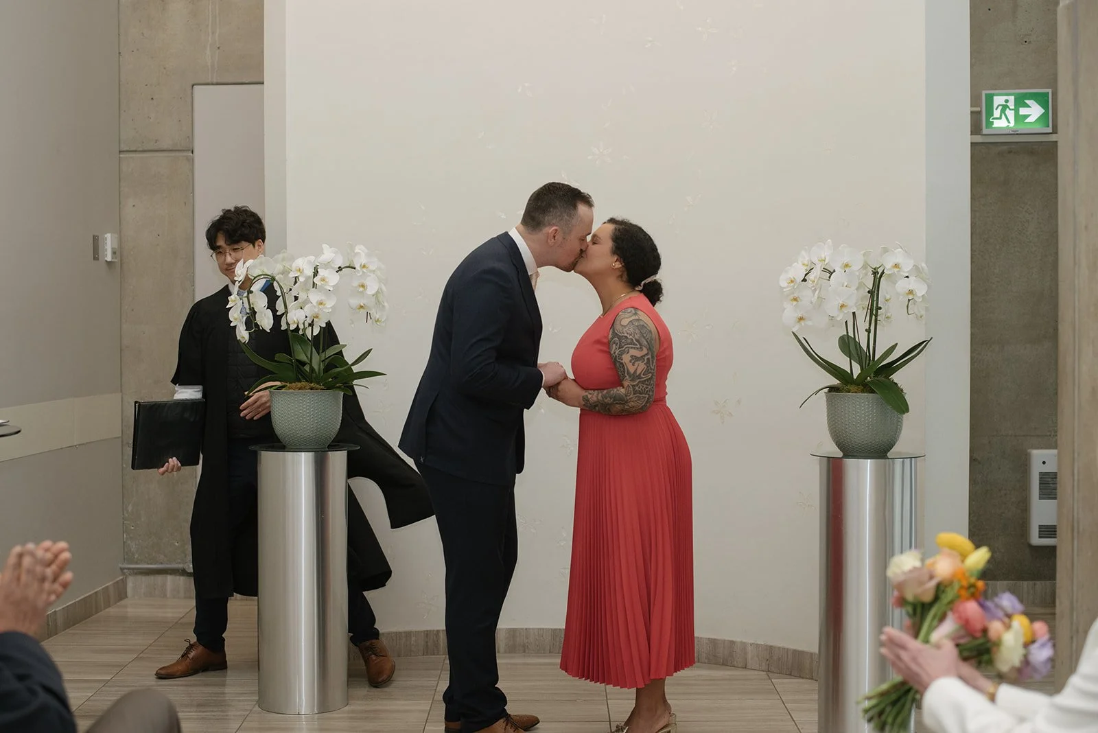 A couple kissing at a wedding ceremony, holding hands, with a man in a suit and a woman in a coral dress with tattoos. 
