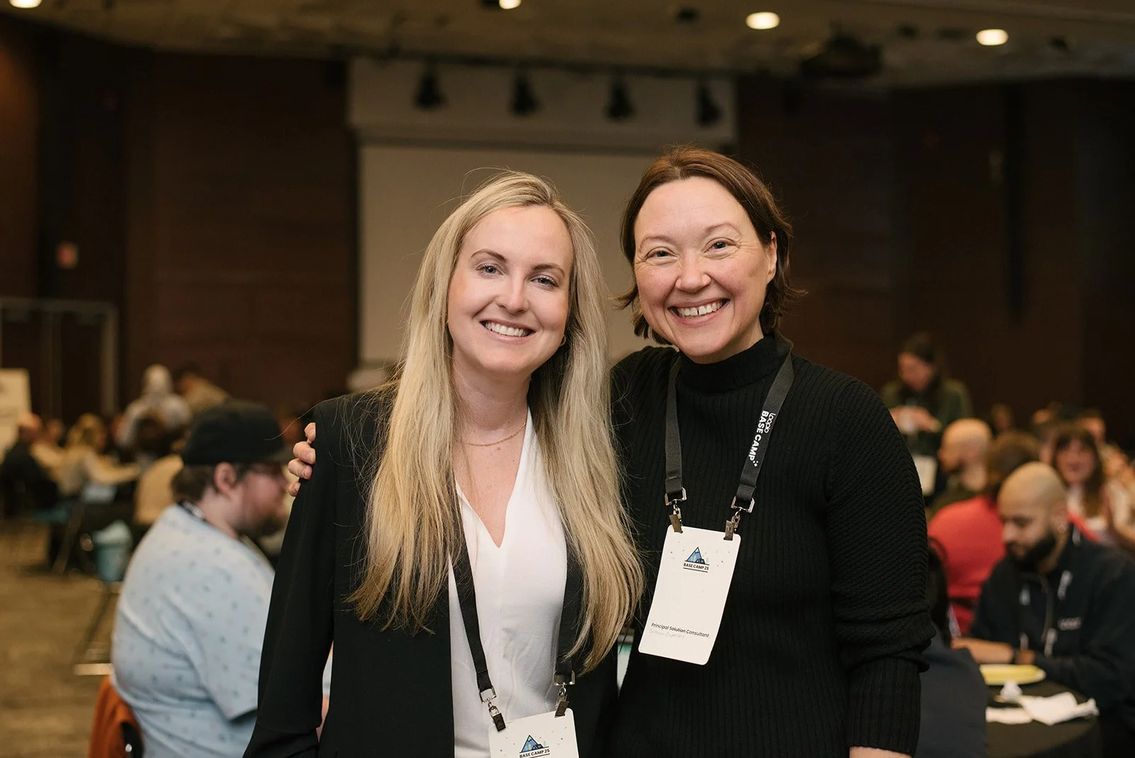 Two women smiling and standing close together at a conference, wearing name tags, with one woman in a black sweater and the other in a black blazer over a white blouse. Conference attendees are in the background.