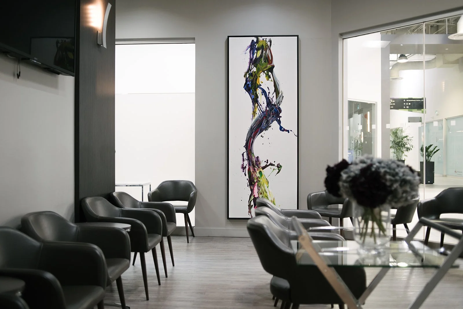 Modern waiting area with black leather chairs arranged around a glass table, a large abstract painting on the wall, and a vase of dark and white flowers on the table.