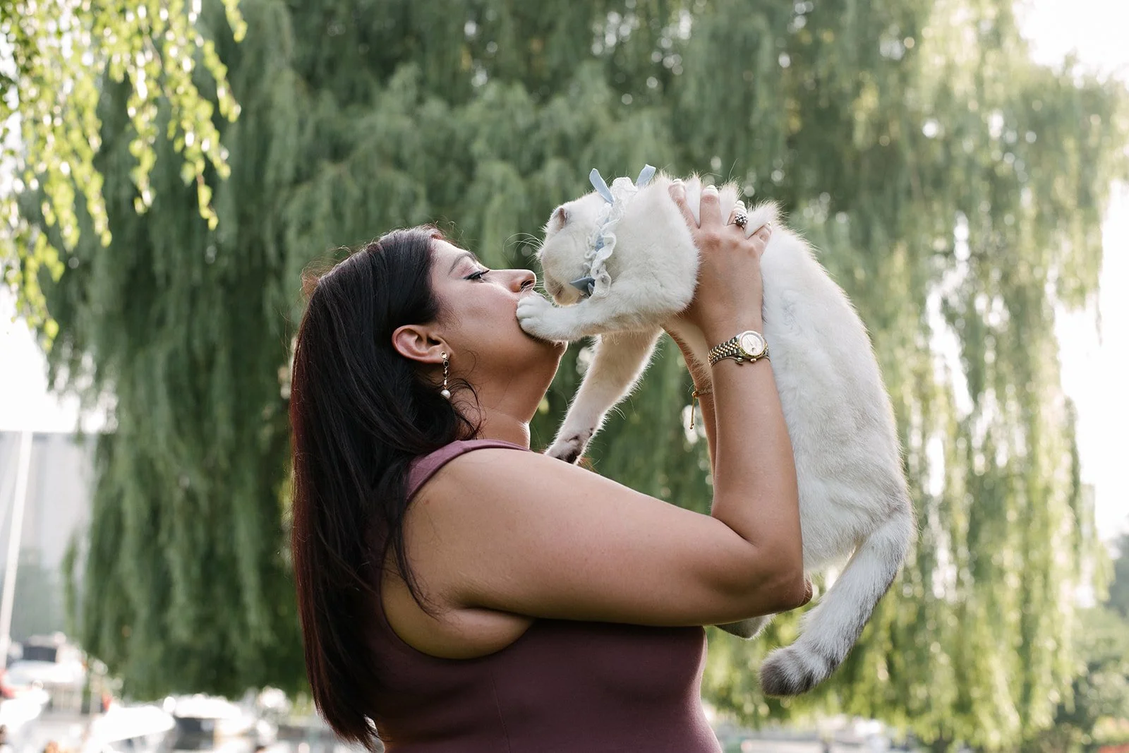 Woman holding up a white scottish fold outside at The Toronto Music Garden with trees in the background.
