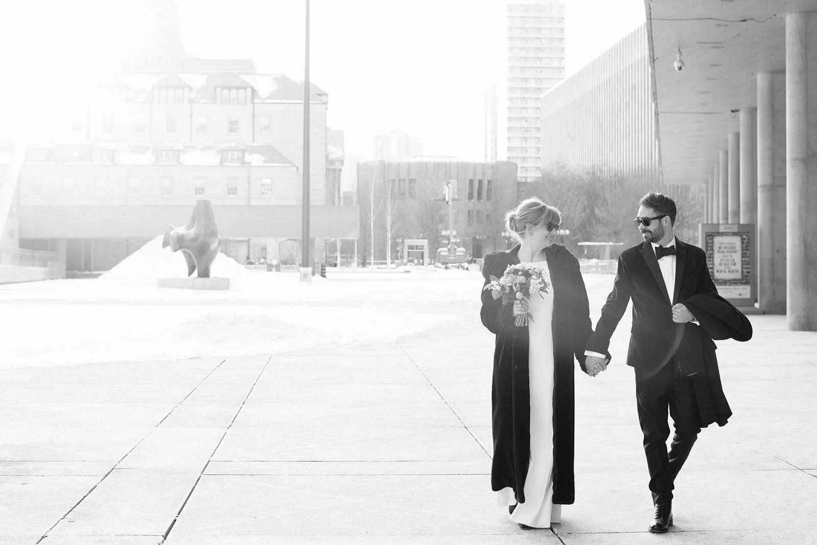 A couple in wedding attire walking outside in a cityscape, holding hands, with the woman holding a bouquet of flowers. The man is wearing a tuxedo, and the woman is wearing a white dress with a dark coat.