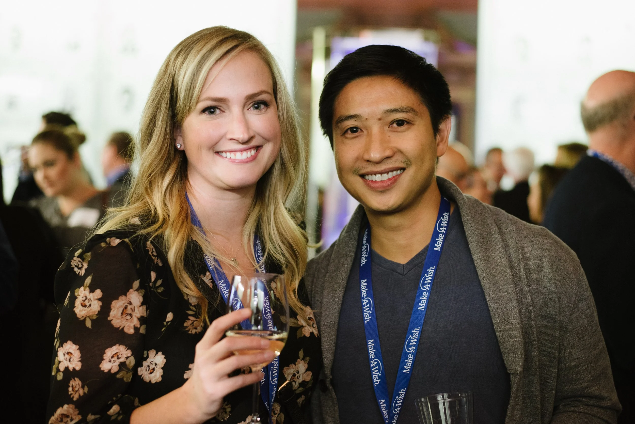A smiling woman with blonde hair holding a drink and a man with dark hair smiling at the camera, both wearing lanyards with 'Make-A-Wish' logo at a social event.