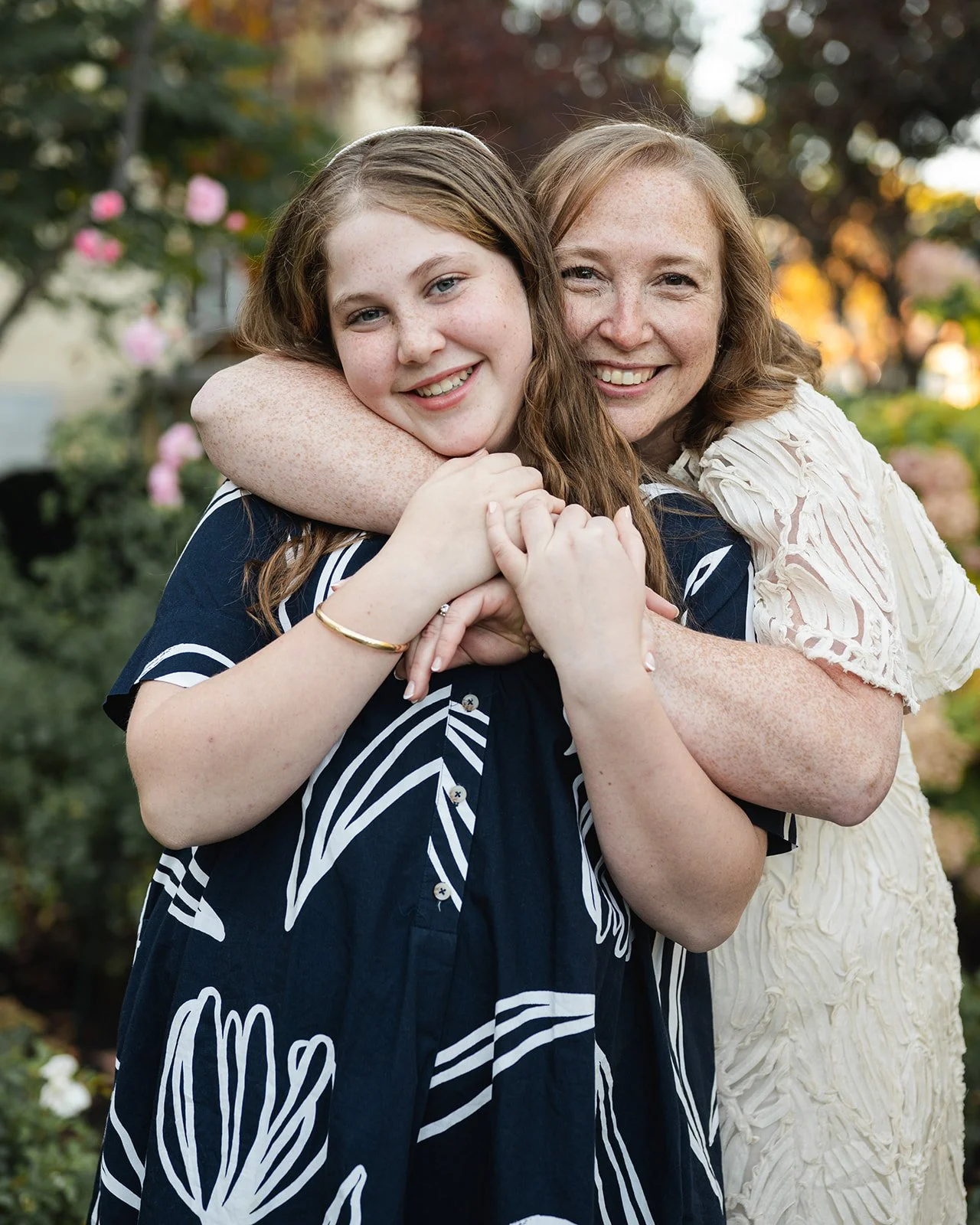 A woman and a girl hugging outside with trees and flowers in the background at Beth Tzedec Congregation.