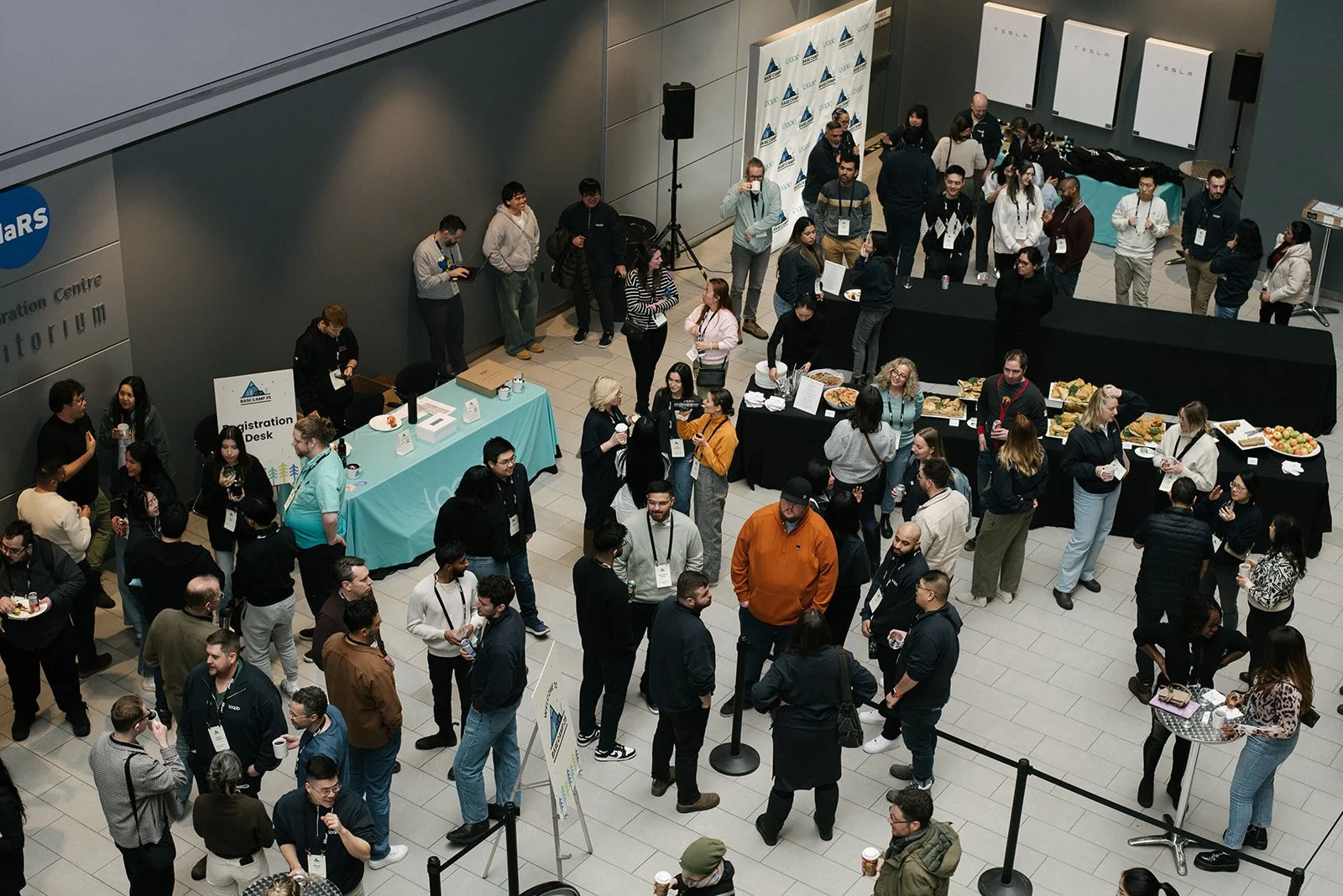 A busy indoor conference registration and refreshment area with numerous attendees socializing, collecting drinks and food, and lining up at registration desks.