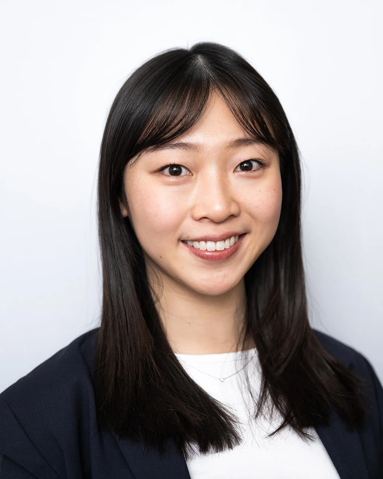 A young woman with shoulder-length dark hair, smiling, wearing a navy blazer over a white top, against a plain white background.