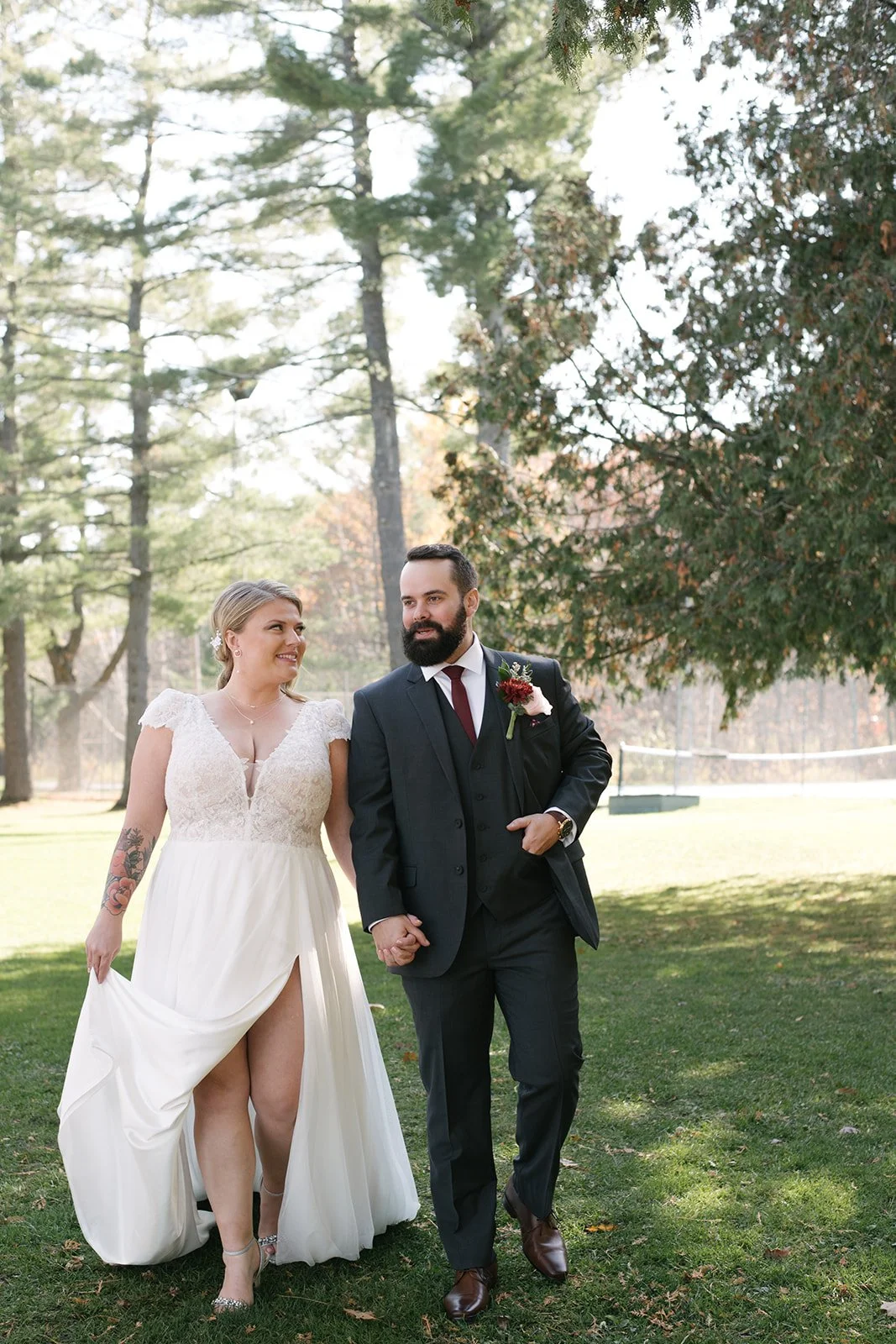 A bride and groom walking hand in hand outdoors during their wedding celebration in a park, with trees and green grass in the background.