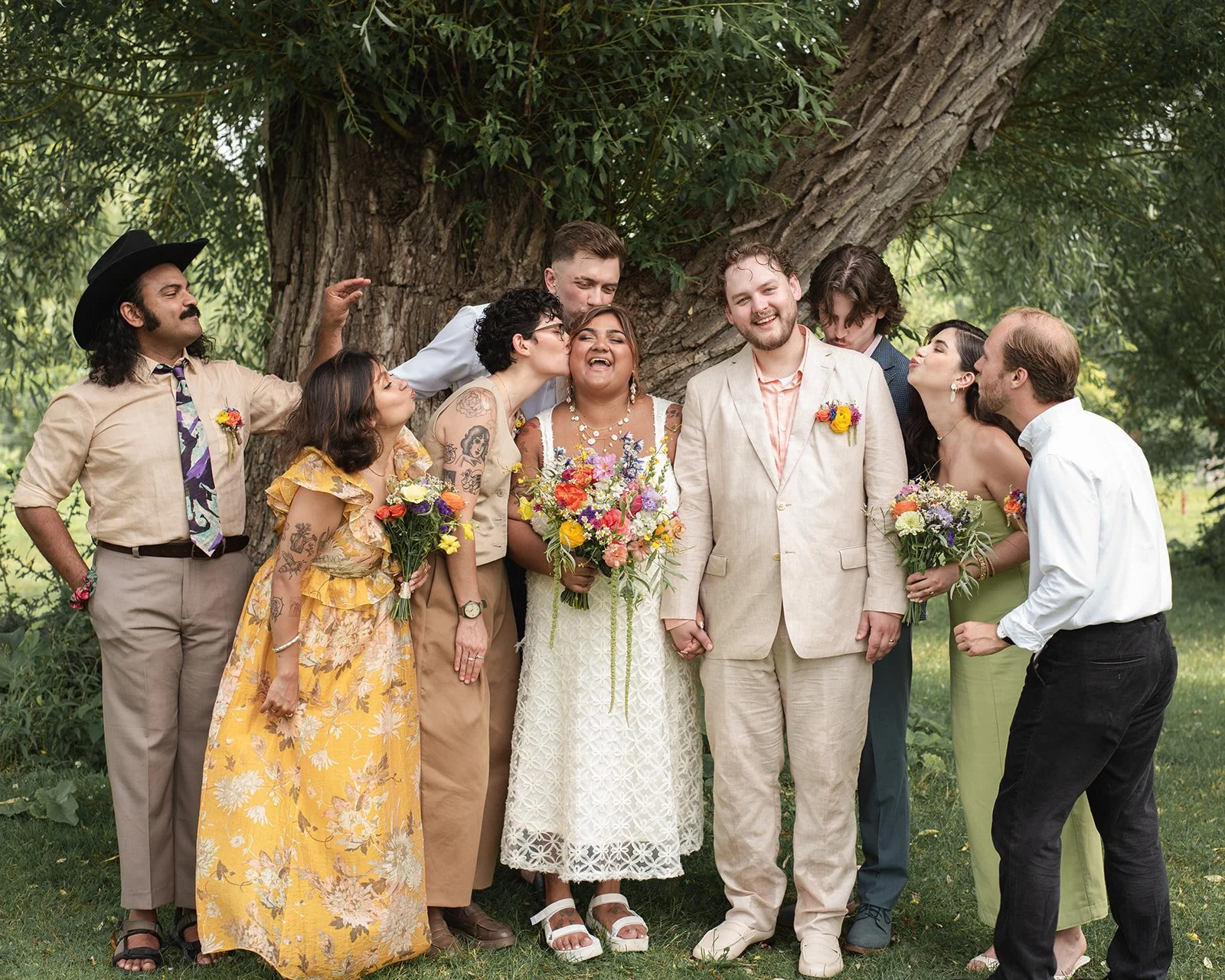 A wedding party of ten people, including the bride and groom, standing outdoors in front of a large tree, all smiling and celebrating with colorful bouquets of flowers.