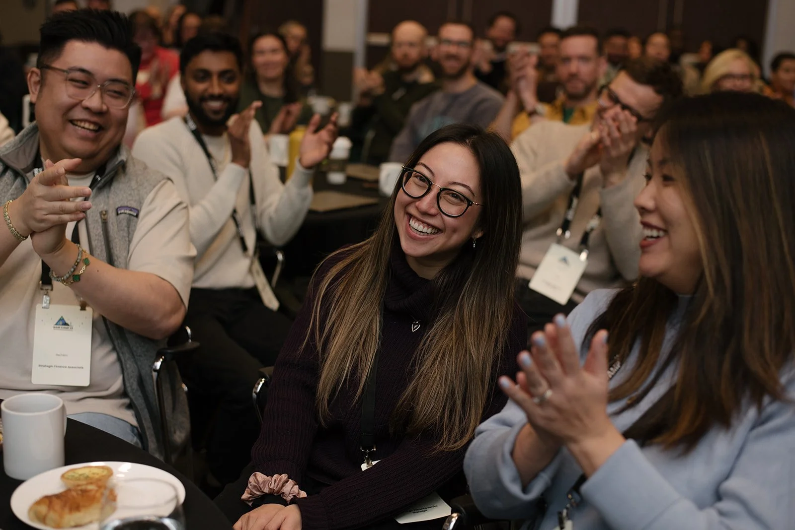 A group of people attending an event, smiling and clapping, sitting around tables with food and drinks.