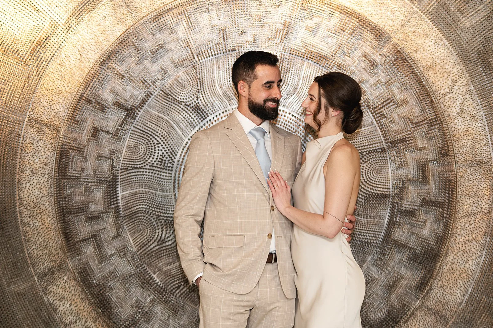A smiling couple dressed in wedding attire standing close together in front of a textured, circular, art installation at Toronto City Hall. The woman is touching the man's chest and they are looking into each other's eyes.