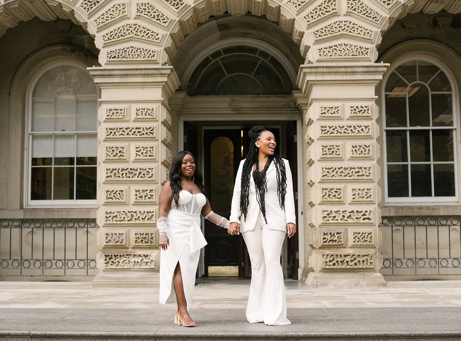 Two women in white wedding attire walking hand in hand in front of Osgoode Hall with arched windows and ornate stonework. They are smiling and holding hands, one with long dark hair and the other with long braided hair.