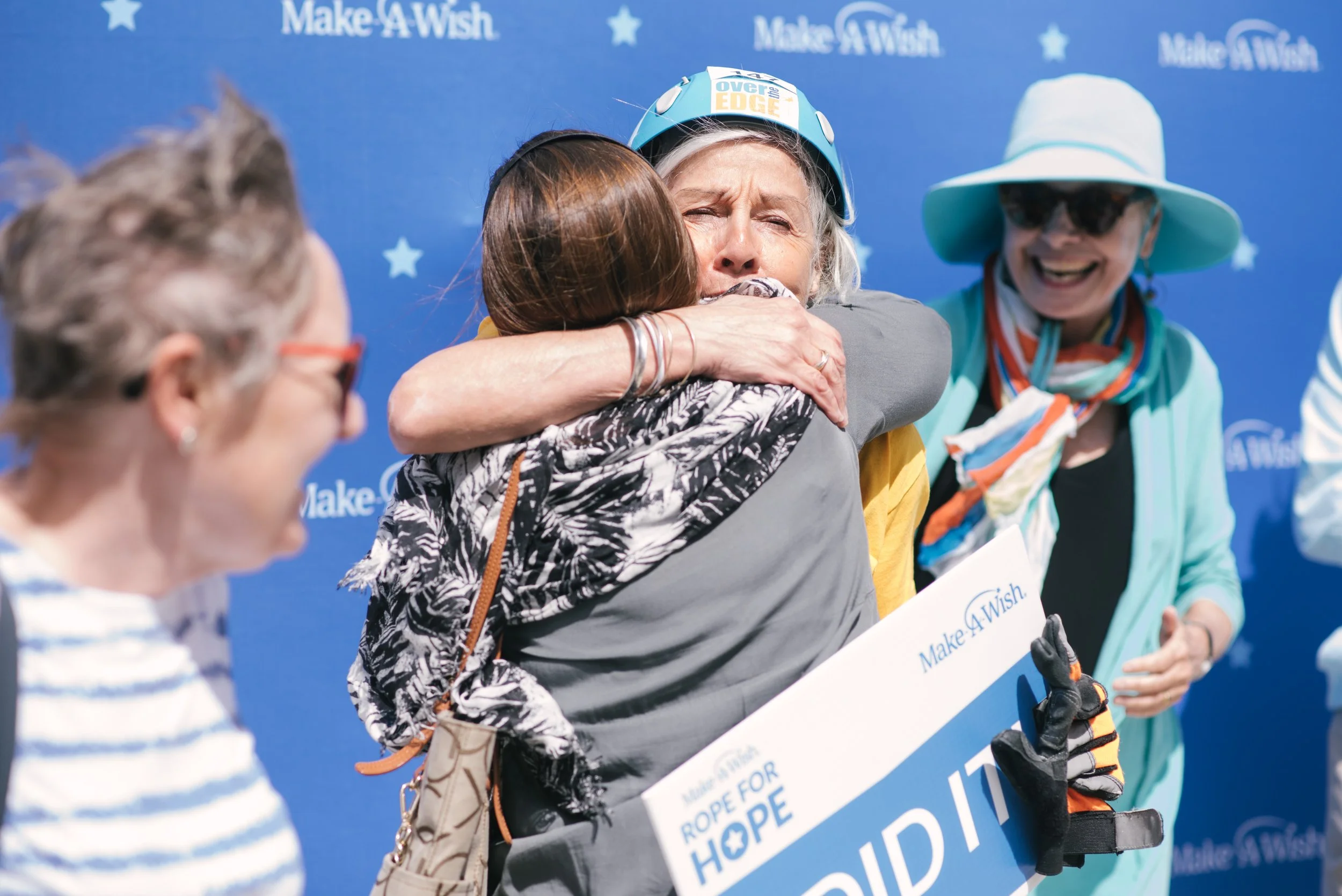Two women hugging in front of a blue background with Make-A-Wish logos, one woman holding a large check for 'Rope for Hope' event.
