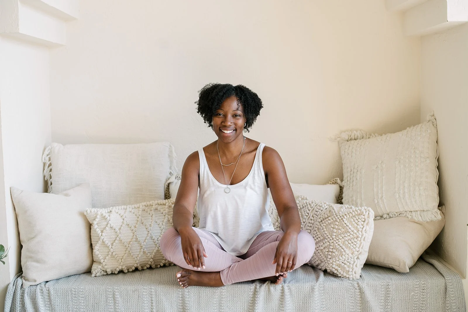 A woman sitting cross-legged on a sofa with beige pillows, smiling at the camera in a cozy, well-lit room inside Mint Room Studios in Toronto.