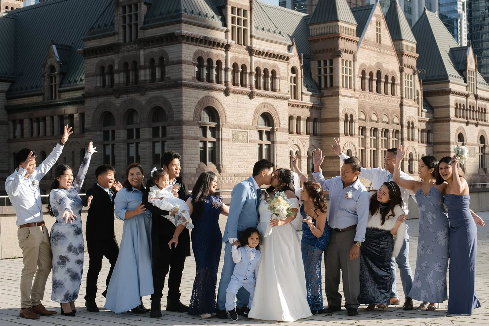 A family portrait taking place after a wedding in front of Old City Hall with a brown stone facade. The bride and groom are kissing among family and friends, who are cheering and raising their hands.