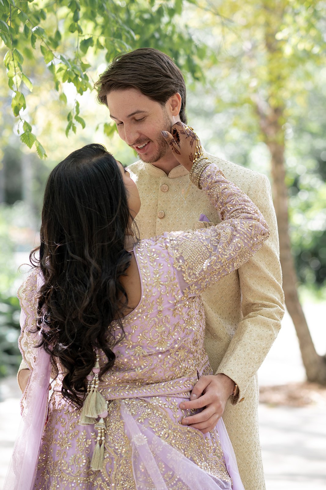 A woman in traditional Indian attire tenderly touches a man in cream-colored formal wear, both smiling and looking at each other outdoors under green trees.