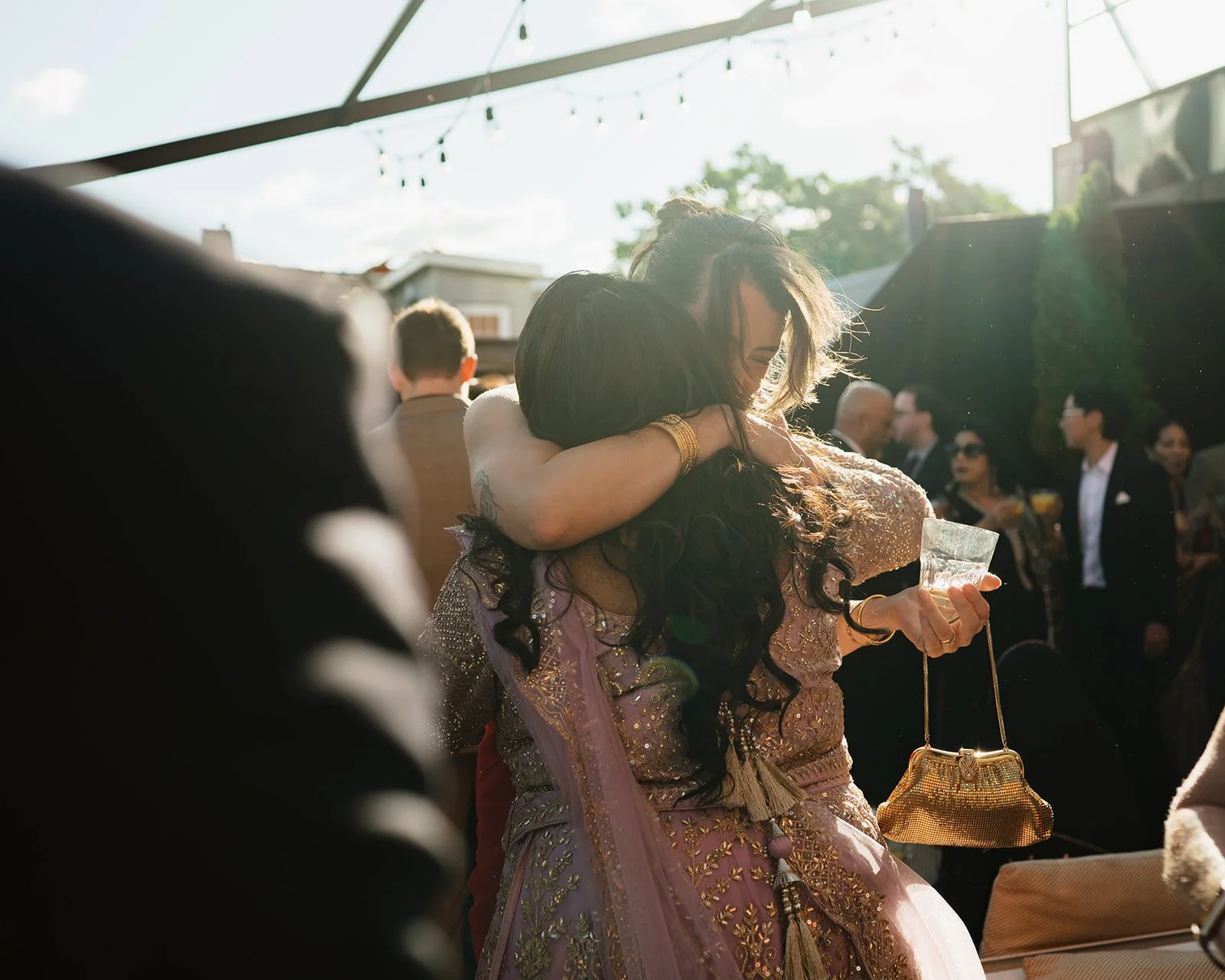 Two women hugging at The Broadview Hotel during cocktail hour at a wedding, one holding a drink and a handbag, with several people in the background.