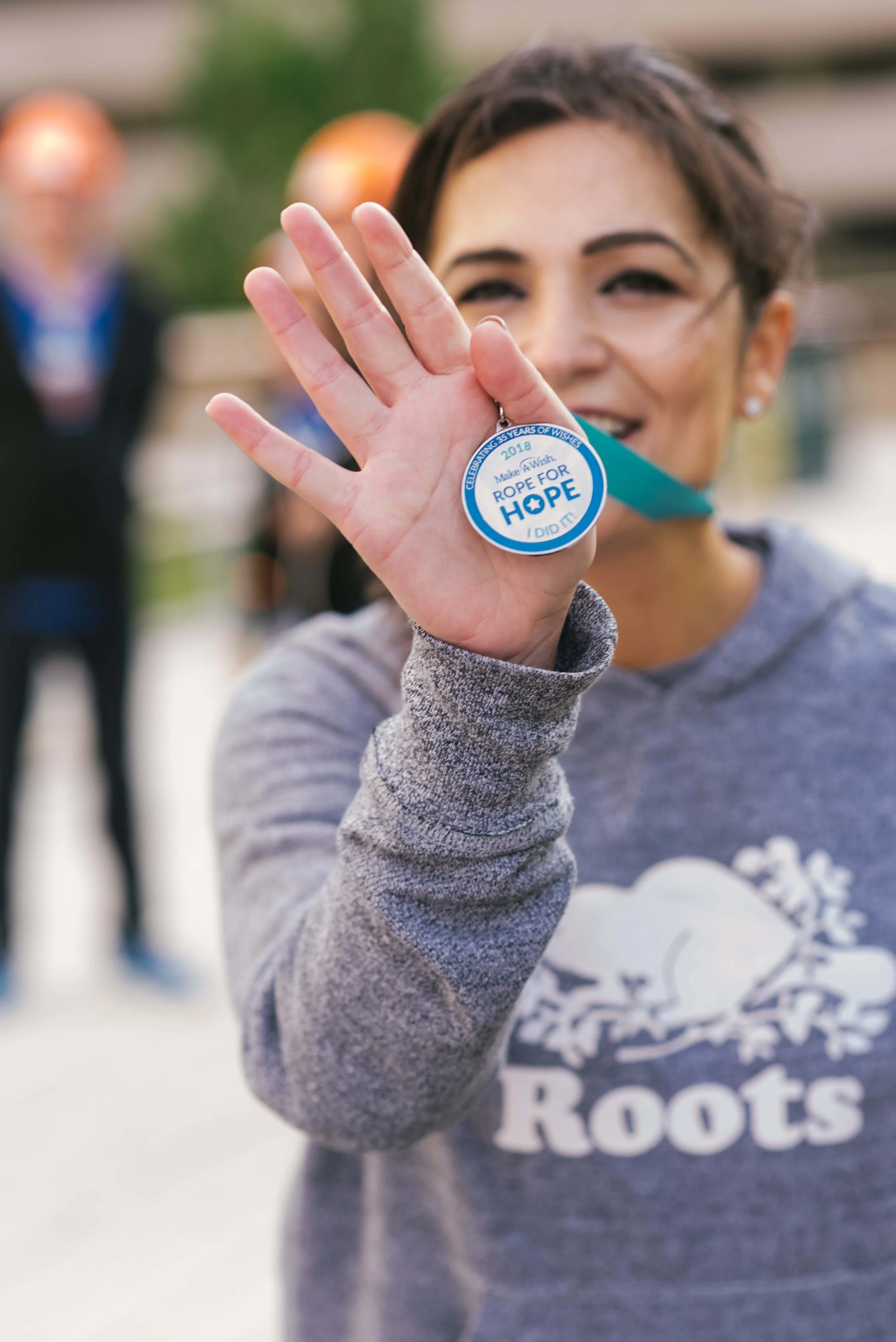 Woman holding a blue and white medal that says "Make A Wish Rope For Hope I Did It." She is smiling and wearing a gray Roots sweatshirt, with people in the background at an outdoor event at Toronto City Hall.